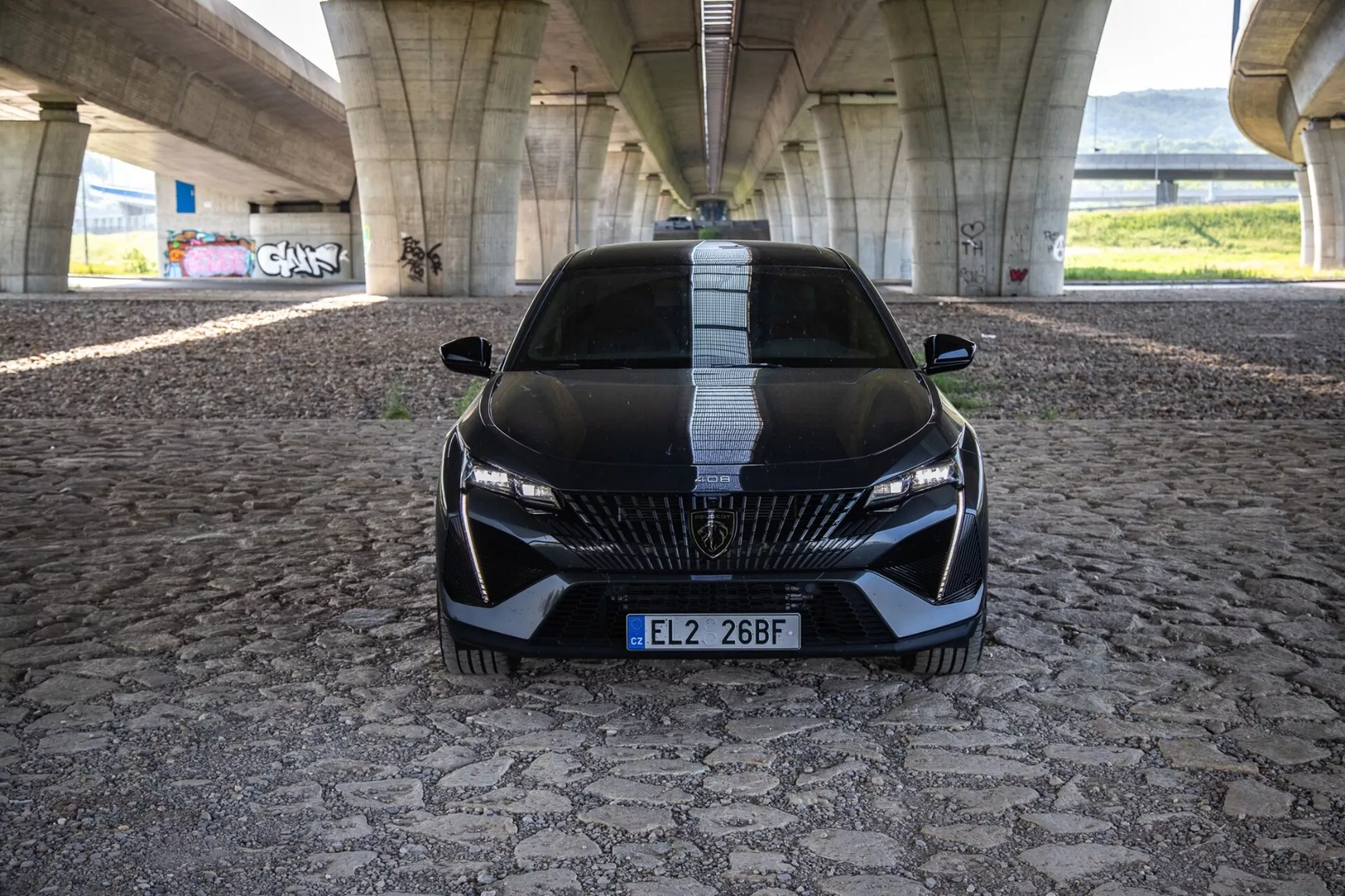 Peugeot 408 2023 i en dynamisk framsyn som visar dess aggressiva ställning och sportiga design med detaljerad grill, slanka LED-strålkastare och aerodynamiska funktioner på ett mönstrat stenläggning under en stadsbro