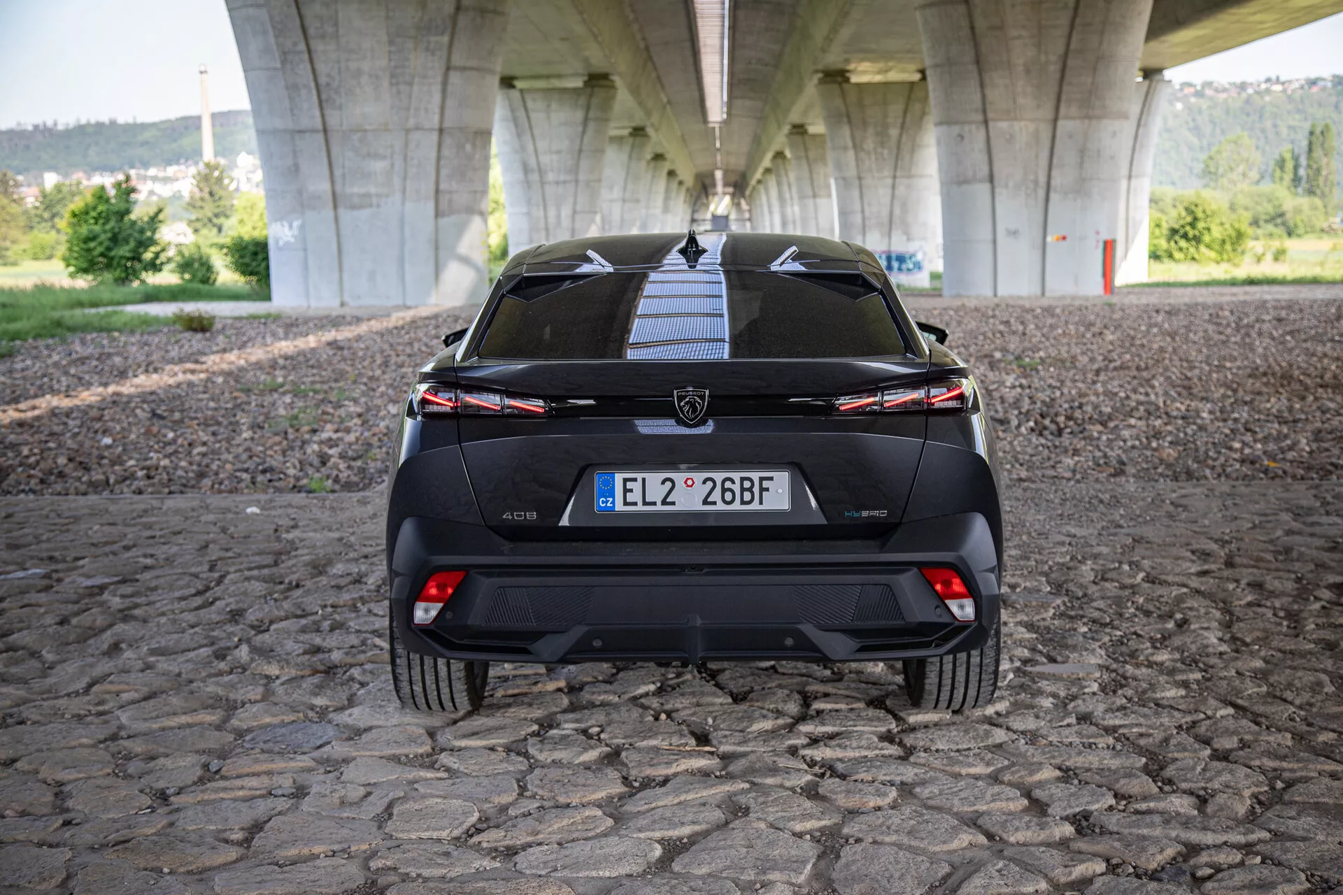 Bakifrån vy av Peugeot 408 2023 som visar baklucka, stötfångare, undre diffusor, bakljus med ett anslutande ljusfält och parkeringsassistanssensorer, fotograferad under en bro på kullerstensbelagd yta