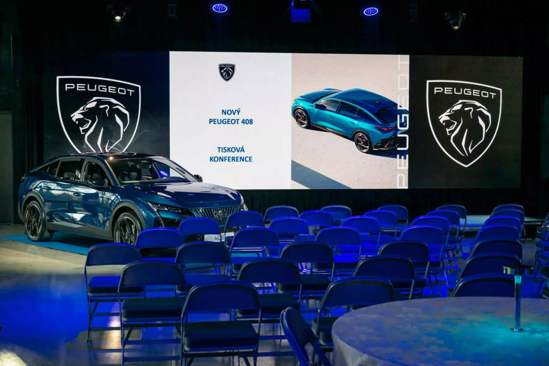 Peugeot 408 visas på en presskonferens, sedd från en 45-graders vinkel som framhäver dess främre och sidoprofiler, slanka design och LED-strålkastare, med digitala skärmar och branding i bakgrunden