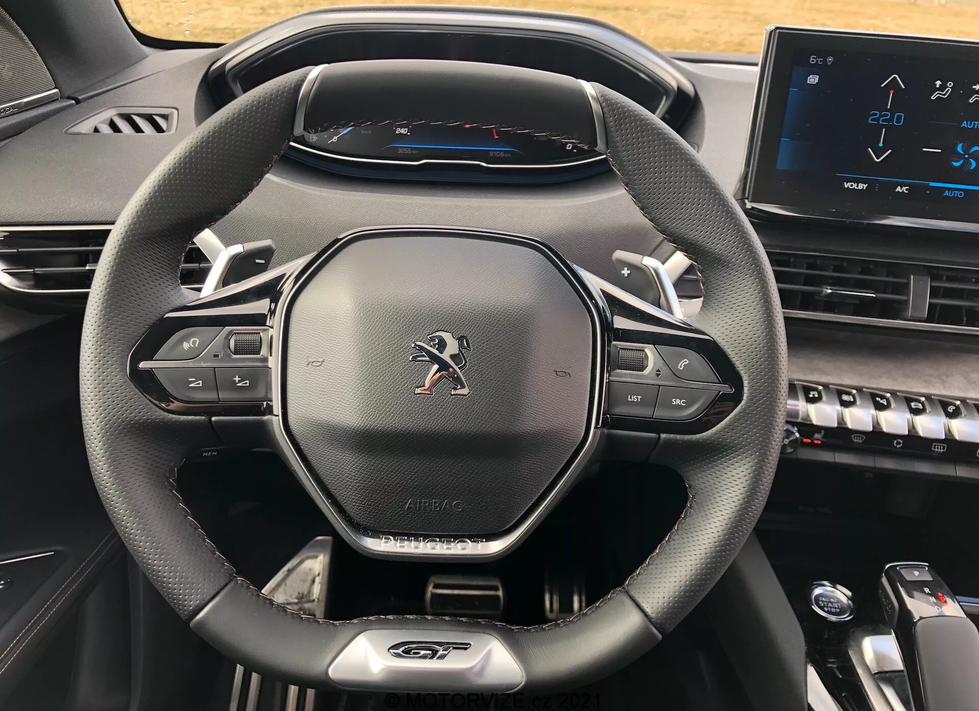 Närbild av förarens cockpit i en Peugeot 5008 från 2021, med fokus på den moderna treekrade läderliknande ratten med multimedia- och farthållarknappar, Peugeot-emblemet, 'AIRBAG'-etiketten, den helt digitala instrumentpanelen som visar en hastighetsmätare med topphastighet på 220 km/h och en del av mittkonsolen med pianoknappar och en elektronisk pekskärm, som visar en samtida och teknologiskt avancerad design.