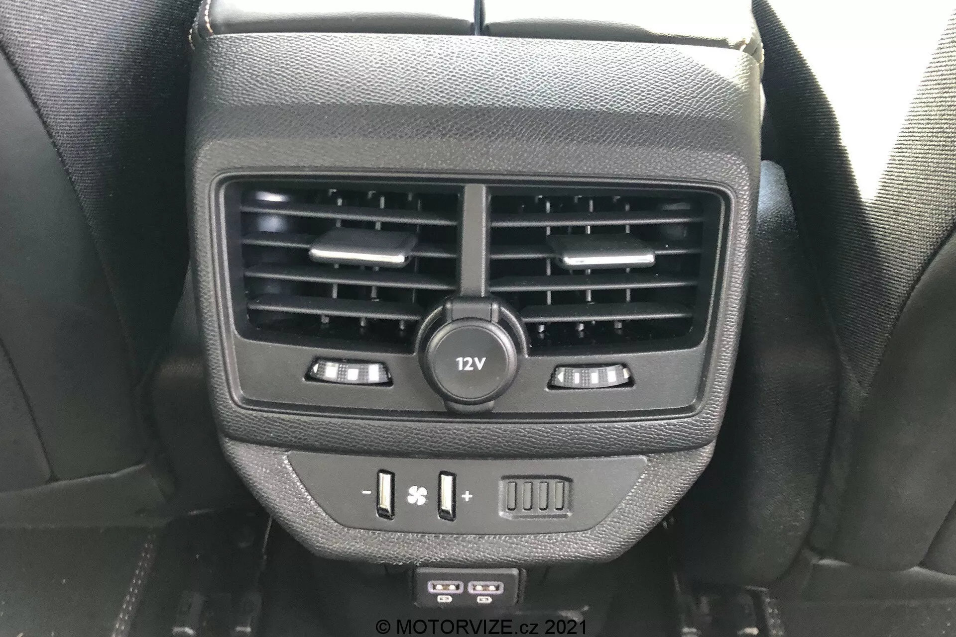 Närbild av funktioner för bakre passagerare på baksidan av mittkonsolen i Peugeot 5008 2021. Bilden visar dubbla luftventiler, ett 12V-uttag, sätesvärmekontroller, USB-portar och en tom knapp, fångad ur en baksätespassagerares perspektiv.