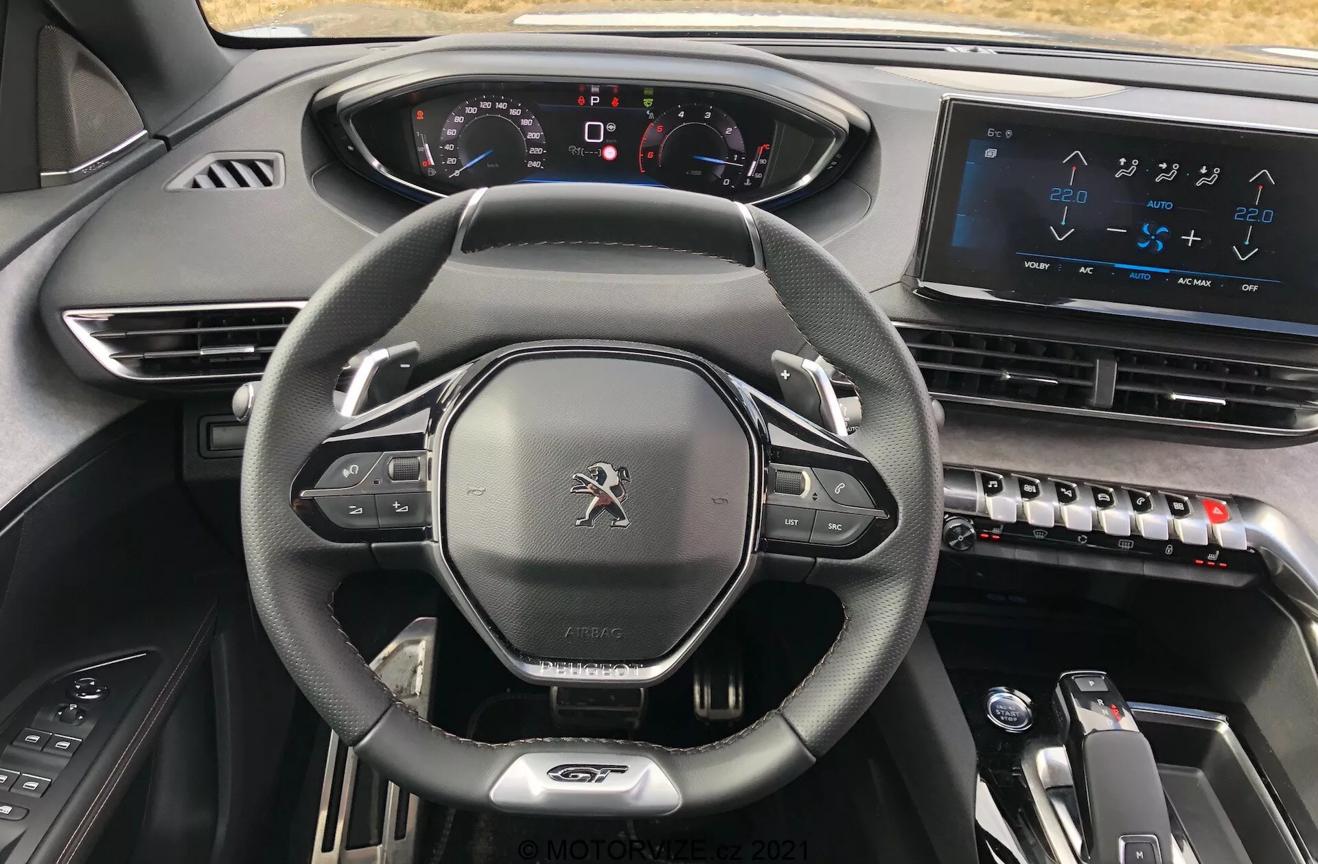 Interiörvy av en Peugeot 5008 2021 från förarsätet, som framhäver den läderklädda ratten med platt botten och inbyggda kontroller, en helt digital instrumentpanel med upplysta indikatorer, en stor pekskärm för infotainmentsystemet på mittkonsolen, eleganta klimatanläggningskontroller, elektronisk växelväljare för automatisk växellåda och en modern och premium inredning med mörka material som uttrycker en högteknologisk och exklusiv estetik.