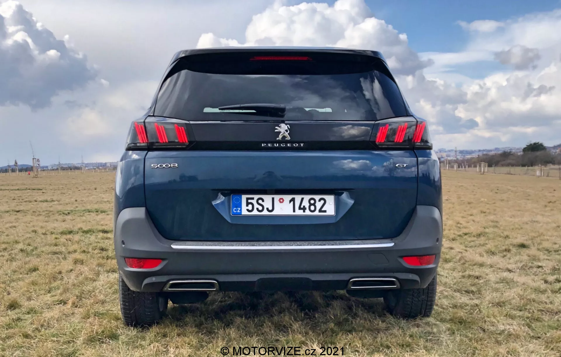 Bakifrånvy av en modern Peugeot 5008 SUV, som framhäver eleganta horisontella bakljus med distinkta kloliknande LED-segment, Peugeot-logotypen och modellbeteckningen '5008'. Mörkblå eller grå färg, parkerad på utomhusterräng med delvis molnig himmel. Synliga detaljer inkluderar bakrutan, bakre vindrutetorkare, högt monterad stopplampa, bagagehandtag och registreringsskylt från Tjeckien med 'CZ'. Reflekterande remsa ovanför stötfångaren med parkeringssensorer och avgasspetsutskärningar är tydliga. Bilden är centrerad med bilen som fyller större delen av ramen.