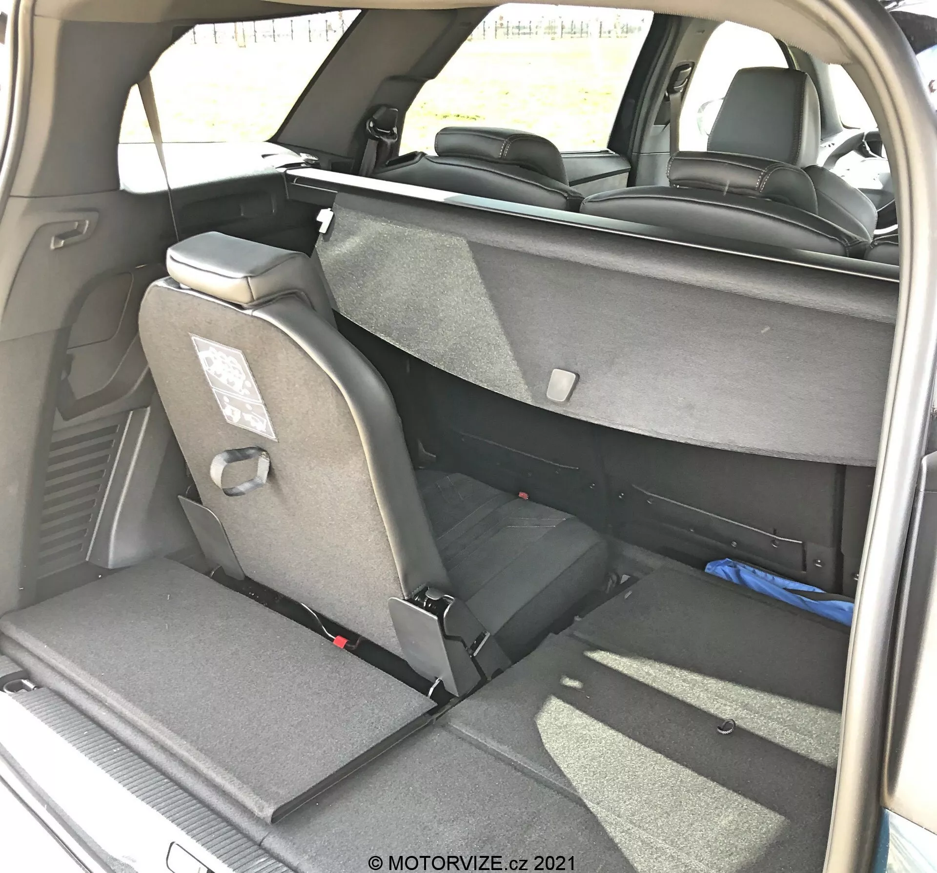 Interiörvy av lastutrymmet i en Peugeot 5008 från 2021 sett från höger bakre trekvarts vinkel, visar en flexibel sittarrangemang med delvis nedfällda baksäten, tygklädsel, nackstöd och säkerhetsbälten. Högra baksätet är helt nedfällt medan vänstra är upprätt, vilket indikerar en delbar 60/40-fällfunktion. Synlig mörkfärgad inredning och mattor med naturligt dagsljus, fotograferad av MOTORVIZE.cz.