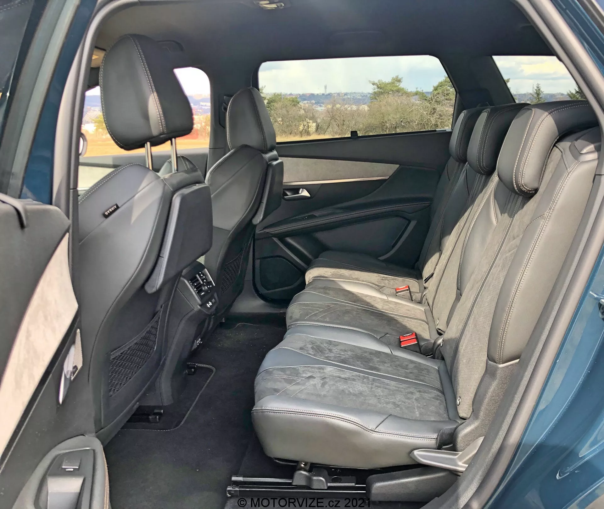 Interiör av en Peugeot 5008 2021 från höger bakre perspektiv som visar det bakre passagerarutrymmet. Den högra bakdörren är öppen och avslöjar dörrpanelens funktioner som handtag, förvaringsfack och högtalare. Tre grå lädersäten med nackstöd är synliga. Mittensätets armstöd är i upprätt läge. Bakre säkerhetsbälten och framsäten med justerbara nackstöd och mittkonsol är också synliga. Baksida kontrollventiler på mittkonsolen, tonat högersidofönster, skymtar av vänster bakdörr som indikerar symmetri. Sammantaget väl underhållen interiör utan synliga tekniska funktioner.