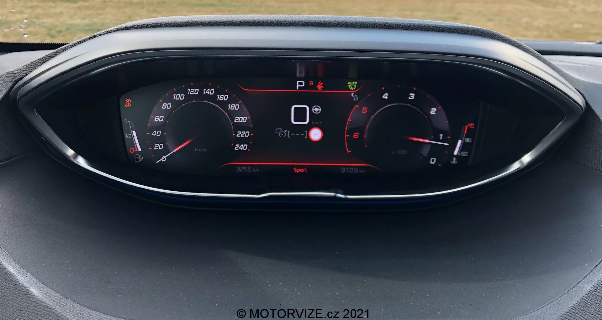 Närbild av instrumentpanelen i en Peugeot 5008 från 2021, med digital display och analoga mätare. Displayen visar hastighetsmätare, varvräknare, växelval och olika fordonsindikatorer såsom säkerhetsvarningar och körläge. Bilen står stilla med körläge 'Sport' aktivt.
