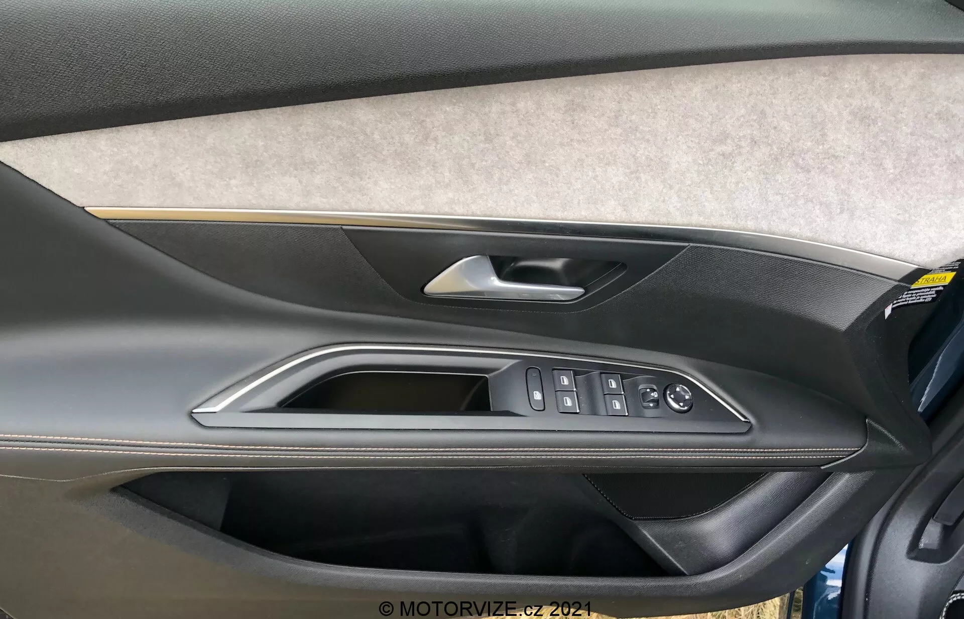 Närbild av interiördörrpanelen på Peugeot 5008 2021 som visar ett sektion klädd i filt eller tyg, en tvåfärgad design i formgjuten plast med en mörkare övre del och en ljusare nedre del, ett högkvalitativt metalliskt eller kromat dörrhandtag, polerad trim och ergonomiska kontroller för elektriska fönster, dörrlås och sidospeglar. En förvaringsficka längst ner på dörrpanelen är också synlig.