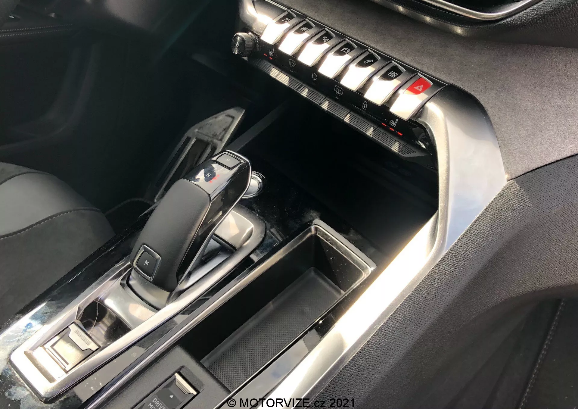Närbild av Peugeot 5008 2021 mittkonsol och främre interiör, med fokus på den automatiska växelspaken och centerdelen. Växelspaken, i 'P'-läge, har en elegant design med glansig svart plast och metalldetaljer. Omkringliggande knappar antyder körlägen och parkeringsbromskontroller. Centerdelen innehåller en rad metallknappar troligen för klimatanläggning, avfrostning och sätesinställningar. Kontrastsöm på läder eller läderimitation och kromdetaljer ökar den lyxiga känslan. Belysning betonar reflekterande ytor och detaljer.