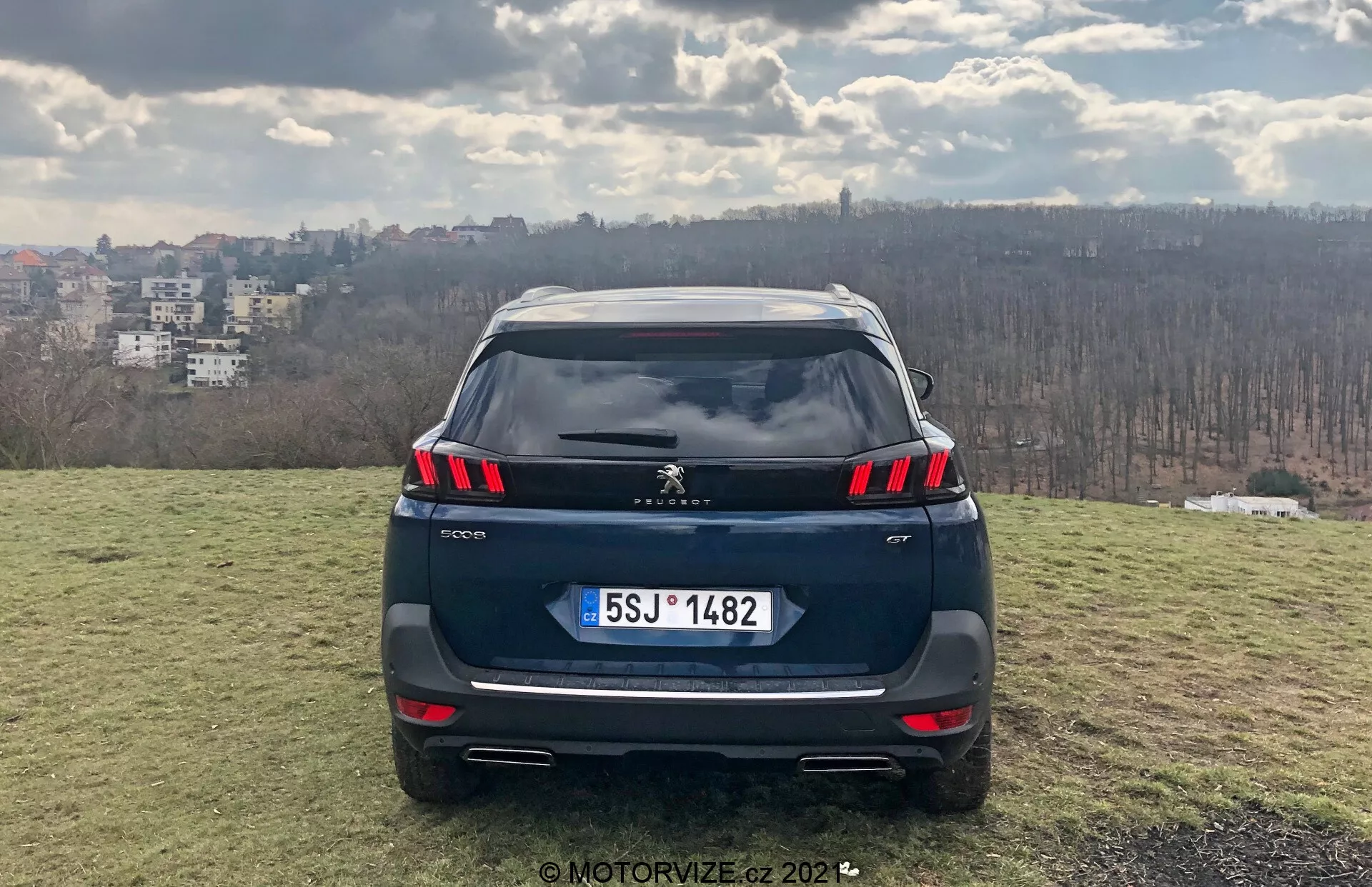 Främre bakre vy av en Peugeot 5008 2021 SUV på en upphöjd gräsbevuxen plats med stadsbild i bakgrunden, med LED-bakljus, integrerade parkeringssensorer i stötfångaren, möjlig backkamera, europeisk stil nummerplåt, dubbla avgasrör, takspoiler och hajfensantenn; daginställning som antyder delvis molnighet, fotograferad av MotorVize.cz 2022.