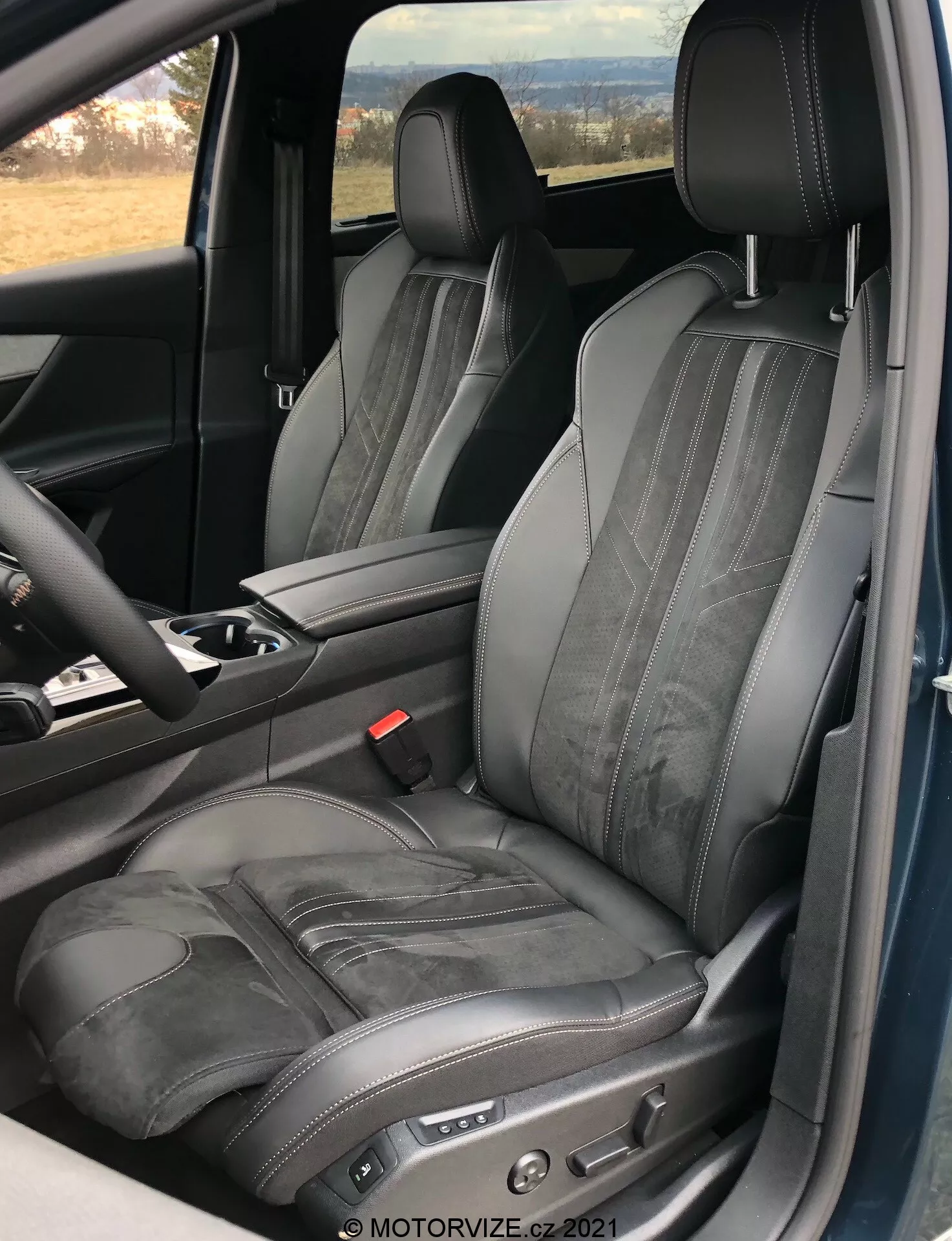 Inre vy av Peugeot 5008 2021 som fokuserar på förarsätet, som visar en justerbar svart lädersits med kontrastsömmar och horisontell präglin, tagen från en öppen dörrperspektiv, som framhäver lyx och ergonomisk design med elektroniska sittjusteringskontroller och en del av mittkonsolen och ratten synlig.