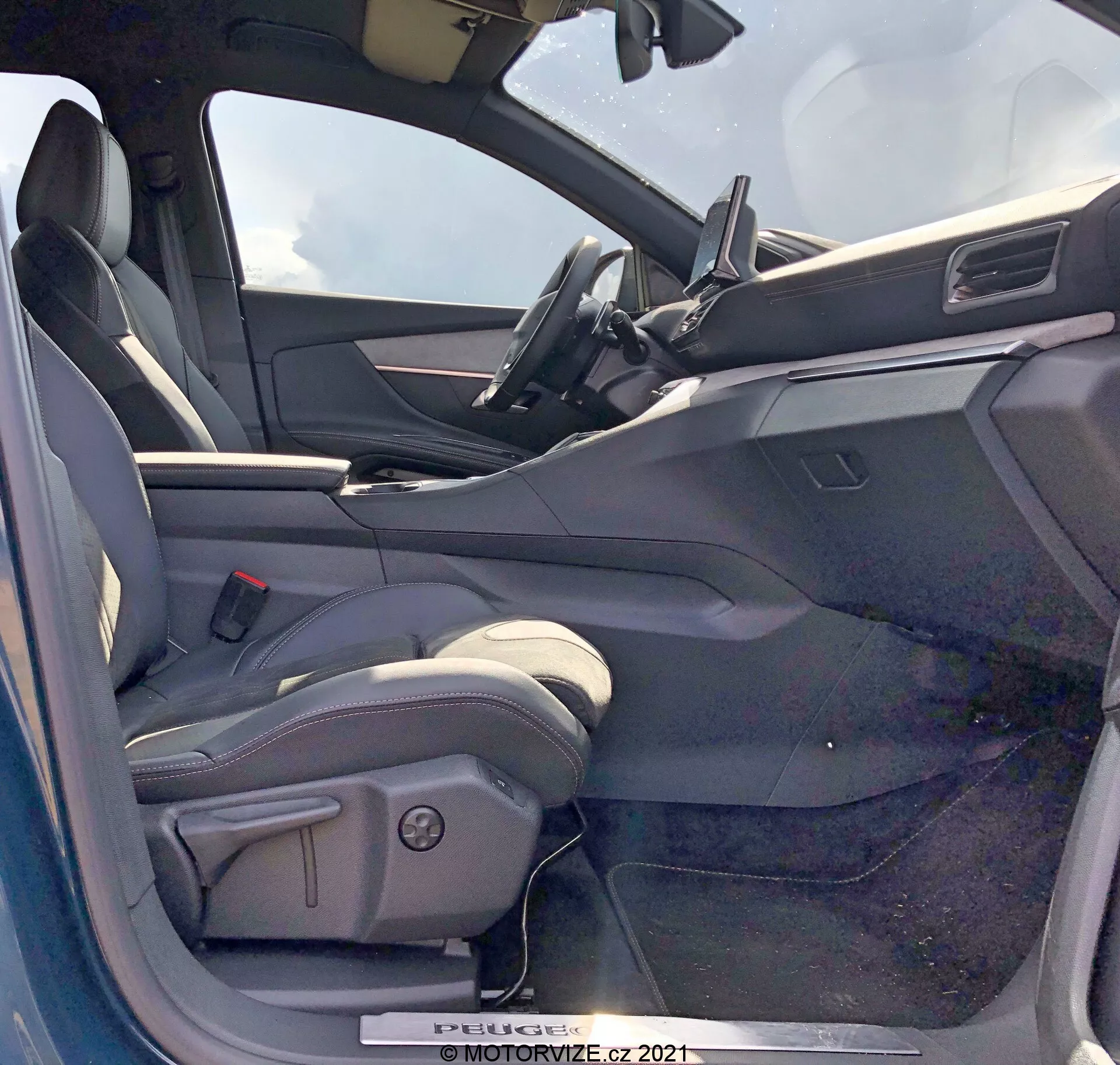 Interiörvy av en 2021 Peugeot 5008 från förarsidan med öppen dörr. Förarsätet är av bucket-stil, troligtvis klätt i läder eller läderliknande material, med sidostöd och elektriska justeringskontroller. Den öppna förarsidans dörr visar en innerpanel med elhisskontroller, eventuellt spegelkontroller och dörrlåsmekanismer. Instrumentbrädan inkluderar en centralt placerad pekskärm för infotainment med minimalistisk design. Den övre delen av ratten är synlig och antyder en läderklädd ratt med monterade kontroller. Interiören har ett tvåfärgat färgschema med mörkare ytor upptill och ljusare ytor nedtill.