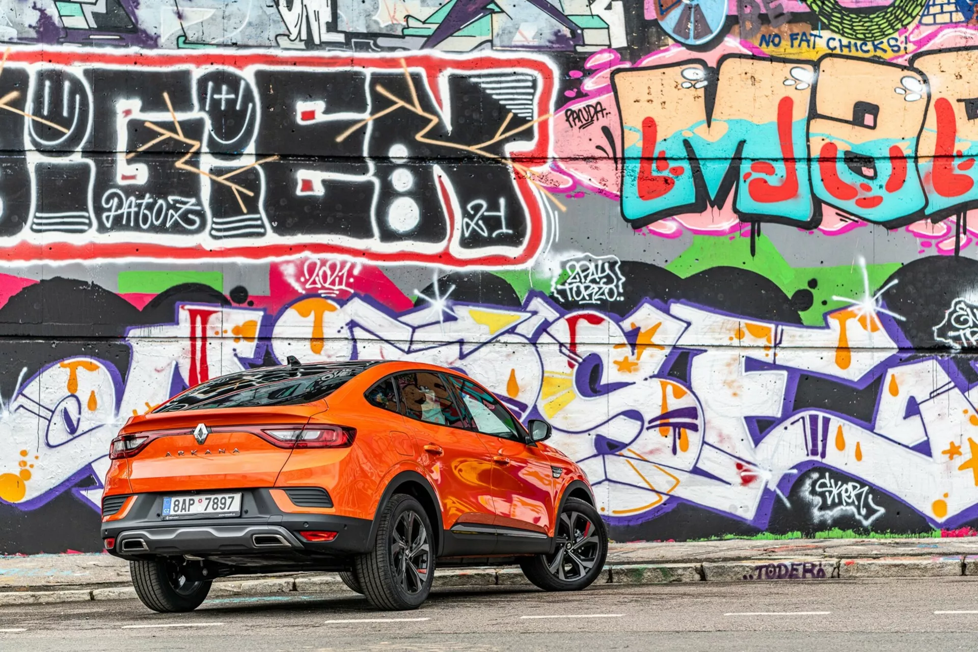 Sido profilvy av en ljusorange Renault Arkana 2021 crossover SUV parkerad på en stads väg. Fotograferad från höger sida med fokus på bakre tre fjärdedelar, fångar bilden bilens sportiga sluttande taklinje, bakspoiler, höga midjelinje, stora stiliga lättmetallfälgar, tonade fönster och aerodynamiska karaktärslinjer. Bakljusen är eleganta och förmodligen LED. Bakgrunden visar en livfull, graffiti-målad vägg, vilket förstärker den urbana estetiken. Fotot, taget i dagsljus, visar mjuka skuggor under bilen, vilket lyfter fram dess designdetaljer.