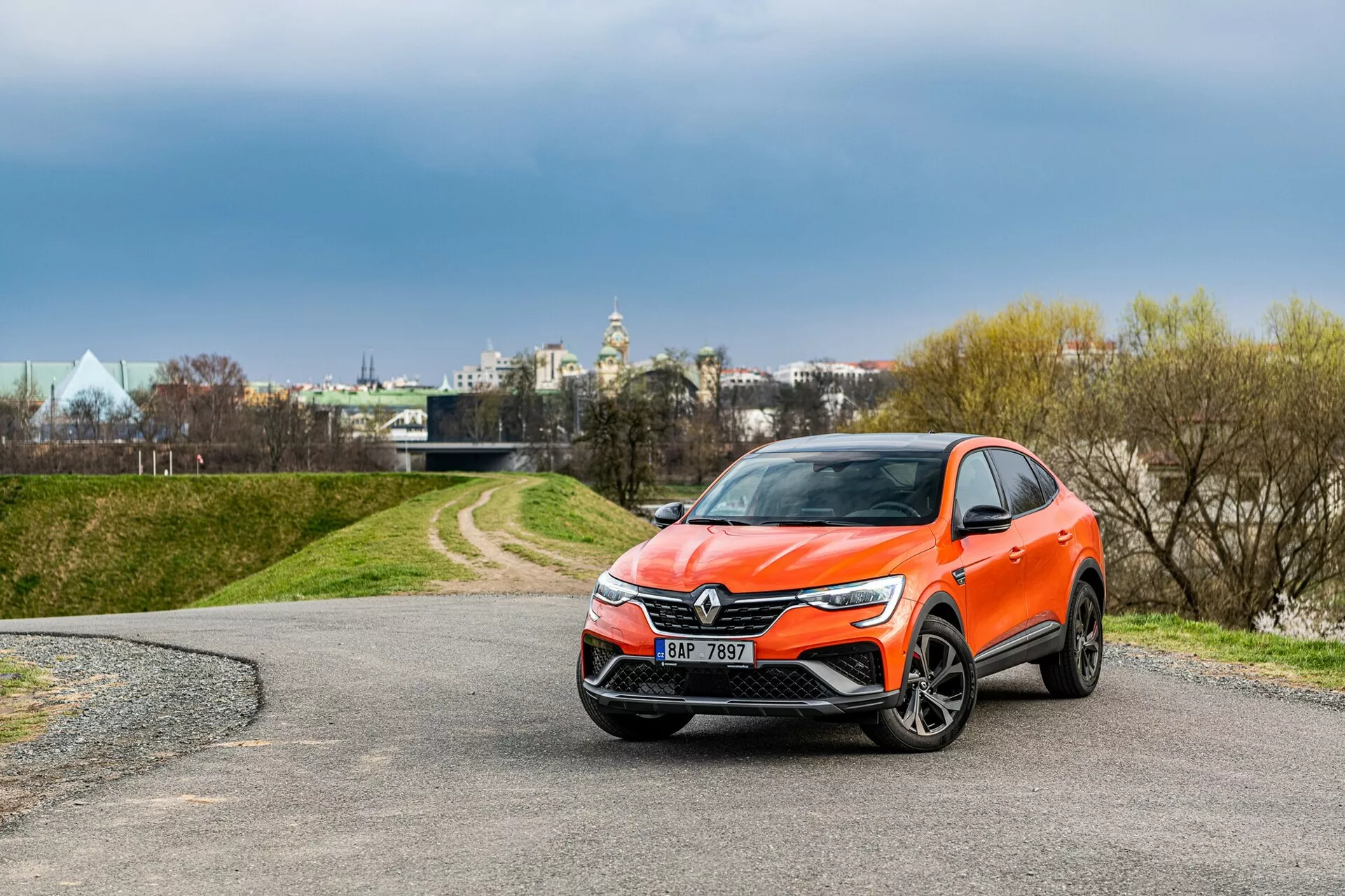 Bilden visar en modern Renault Arkana crossover SUV från 2021 parkerad på en slingrande väg. Fotograferad från trekvarts frontvy, visar bilden tydligt fordonets främre grill med ett framträdande emblem, eleganta strålkastare som sträcker sig mot sidorna och en exakt stylad framstötfångare troligen utrustad med dimljus och sensorer. Aerodynamiskt designade sidospeglar med eventuellt integrerade blinkers, aluminiumfälgar och en levande orange karossfärg förhöjer utseendet. Miljön inkluderar träd och en molnig himmel i bakgrunden, med strukturer som antyder ett stadsområde i fjärran.