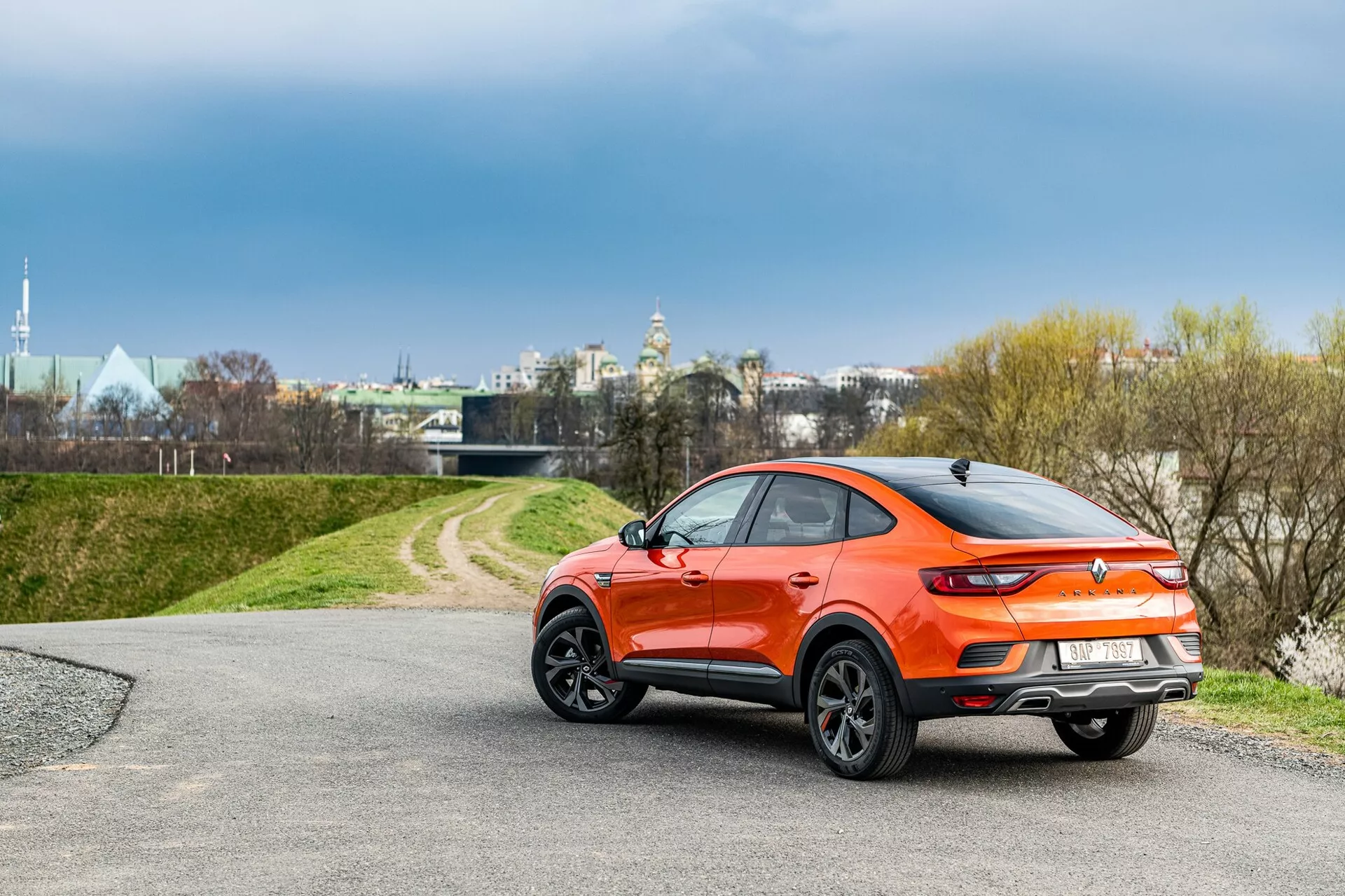 Bakre trekvarts vy av en 2021 Renault Arkana hatchback med en sportig crossoverdesign i en vibrerande orange metallicfärg. Bilen har en sluttande taklinje, svarta plastdetaljer runt hjulhusen, en spoiler ovanpå luckan, eleganta LED- bakljus och stora fälgar med flera ekrar. Den är placerad på en asfalterad väg som leder till ett gräsbevuxet område, under molniga förhållanden nära en urban men lugn miljö.