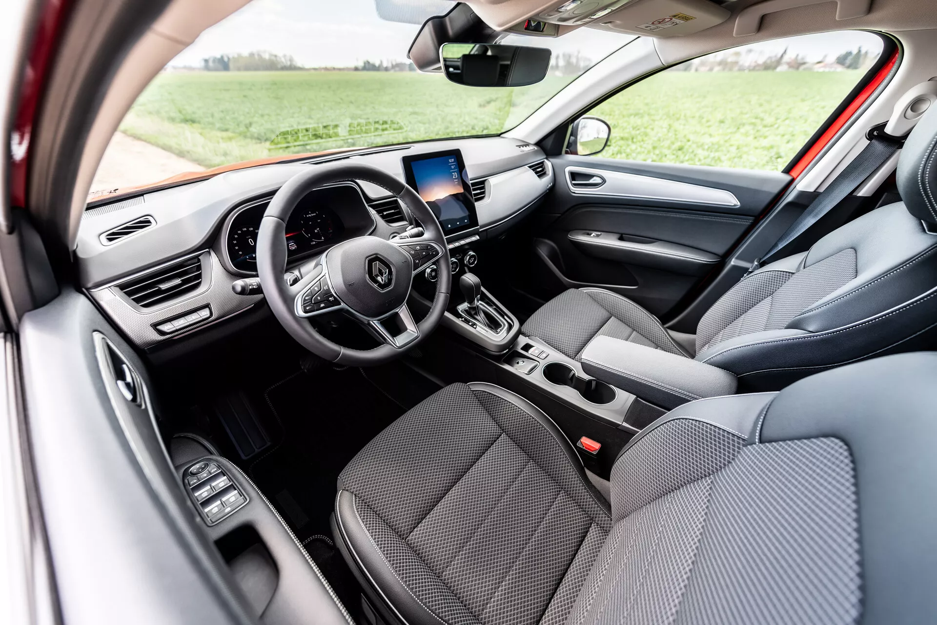 Interiörvy av Renault Arkana 2021 som visar grå tygklädda framsäten, multifunktionsratt med tre ekrar och integrerade kontrollknappar, digital instrumentpanel, infotainmentskärm med GPS-navigering ovanför mittenluftventilerna, manuella kontroller för luftkonditionering, automatväxelspak och kontroller för elektriska fönsterhissar på förarsidan dörr, belyst av naturligt dagsljus i en utomhusmiljö