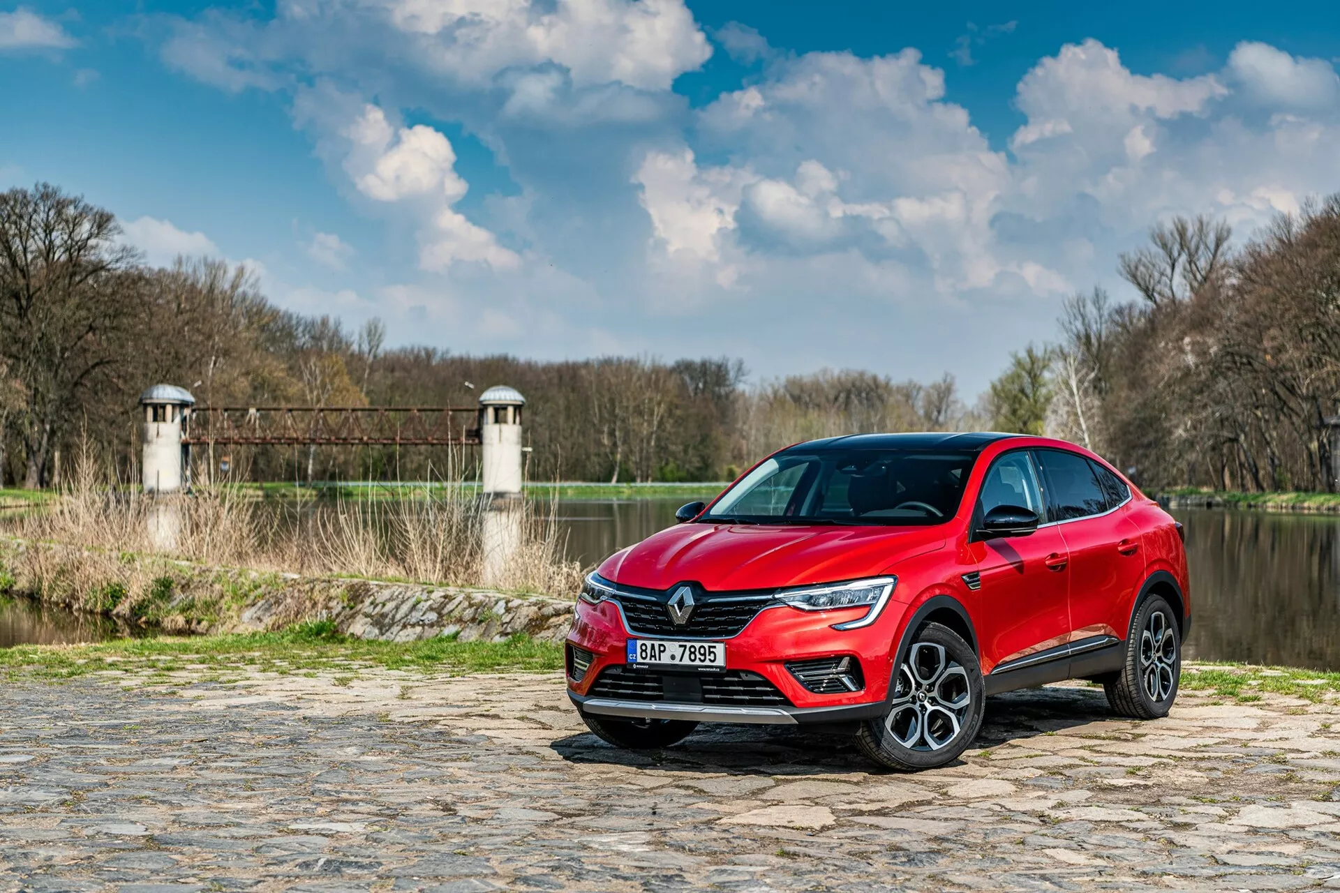 Bilden visar en Renault Arkana 2021 parkerad på en kullerstensväg nära en flod eller sjö, med växtlighet och strukturer som liknar damm- eller brotorn i bakgrunden. Bilen är fångad från en låg vinkel, trekvarts vy framifrån, vilket framhäver frontgrillen, LED-strålkastarna och en del av sidoprofilen inklusive förardörren och framskärmen. De moderna designelementen, såsom den skarpa LED-ljussignaturen och de stora lättmetallhjulen, antyder en högre utrustningsnivå. Den reflekterande metalliska lacken ökar den estetiska dragningskraften, medan de möjliga förarassistanssensorerna och teknologin som är inbyggd i stötfångarna och sidospeglarna förbättrar funktionaliteten.