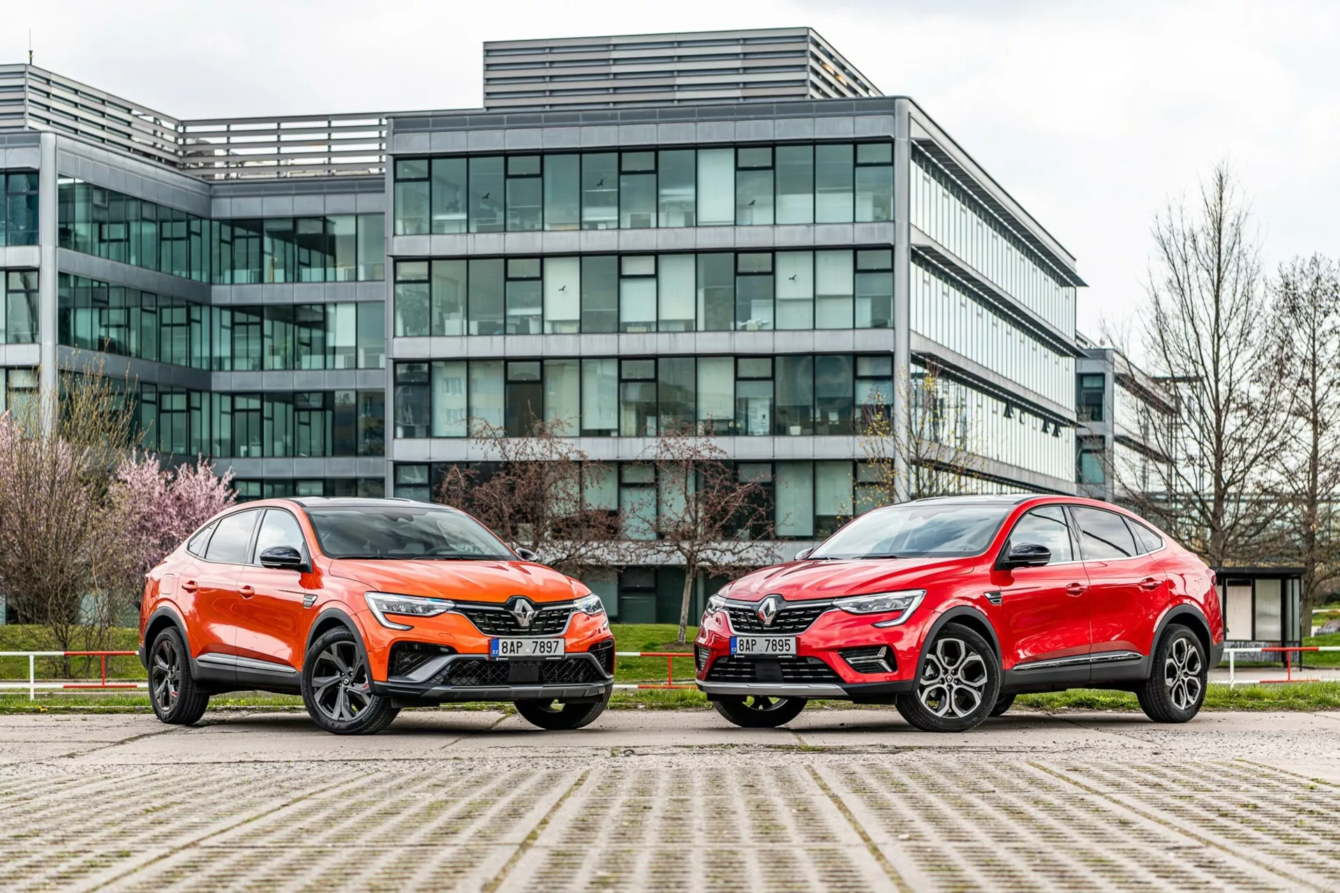 Bilden visar två SUV-bilar parkerade sida vid sida framför en modern kontorsbyggnad med glasfasad. Bilen till vänster är lackerad i en livlig orange färg med metallisk lyster och har ett distinkt frontgaller, skarpa LED-strålkastare, en skulpterad motorhuv och stora fler-ekrade aluminiumfälgar med lågprofildäck. Bilen till höger har en metallisk röd finish, med en liknande övergripande silhuett, skarpa strålkastare, framträdande grill och snygga stora lättmetallfälgar. Båda fordonen sticker ut med sina vinklade linjer, aerodynamiska speglar och kontrasterande underkroppstrim. Fotot, taget från en låg vinkel nära marknivå, framhäver den urbana miljön och betonar fordonens design, närvaro och avancerade tekniska funktioner.