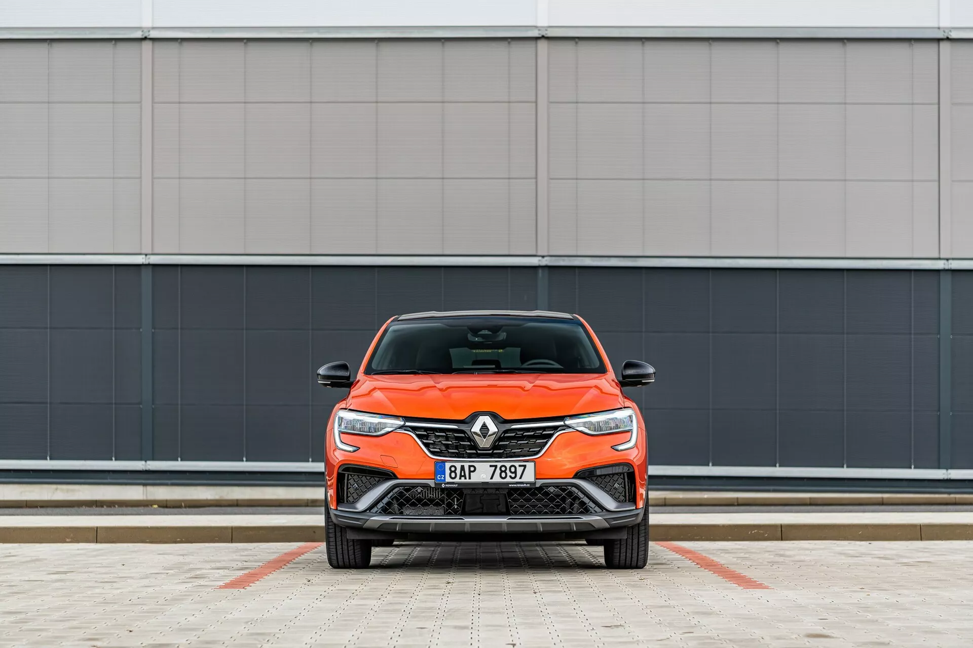 Frontal vy av en ljusorange Renault bil, som framhäver dess moderna design med eleganta strålkastare och frontgrill, parkerad vinkelrätt mot kameran mot en minimalistisk grå vägg på en markerad parkeringsplats.