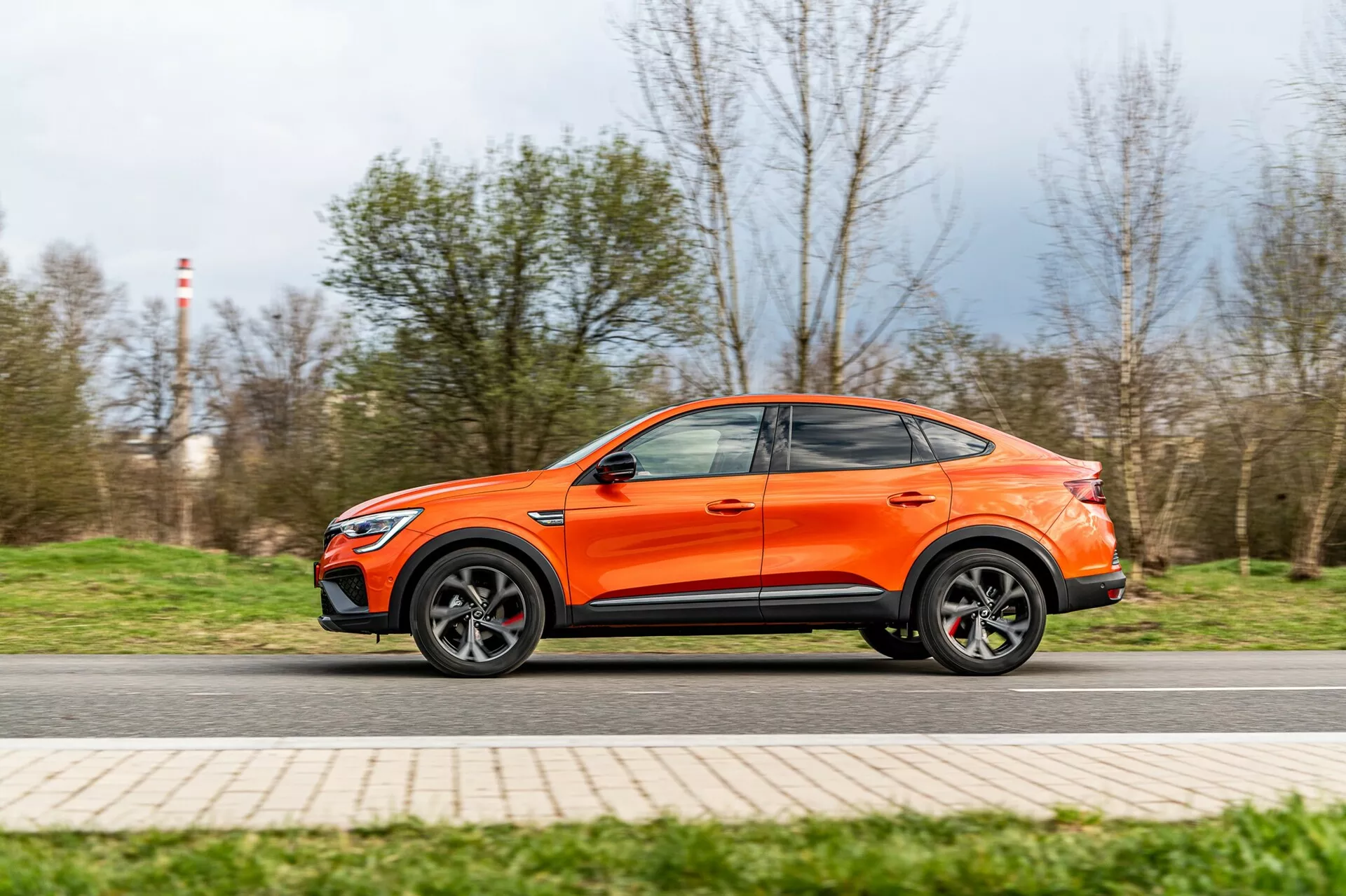 Dynamisk sidoprofilbild av en orange Renault Arkana 2021 crossover SUV i rörelse, tagen från en låg vinkel som betonar markfrigång och sportig hållning. Bilens hela längd är synlig, vilket visar designlinjer, lättmetallfälgar, konturerade sidodörrar och ett kupéliknande tak. En lätt rörelseoskärpa på hjulen och bakgrunden antyder moderat hastighet, med bilen i skarpt fokus. Bilden framhäver fordonets moderna och aerodynamiska design.