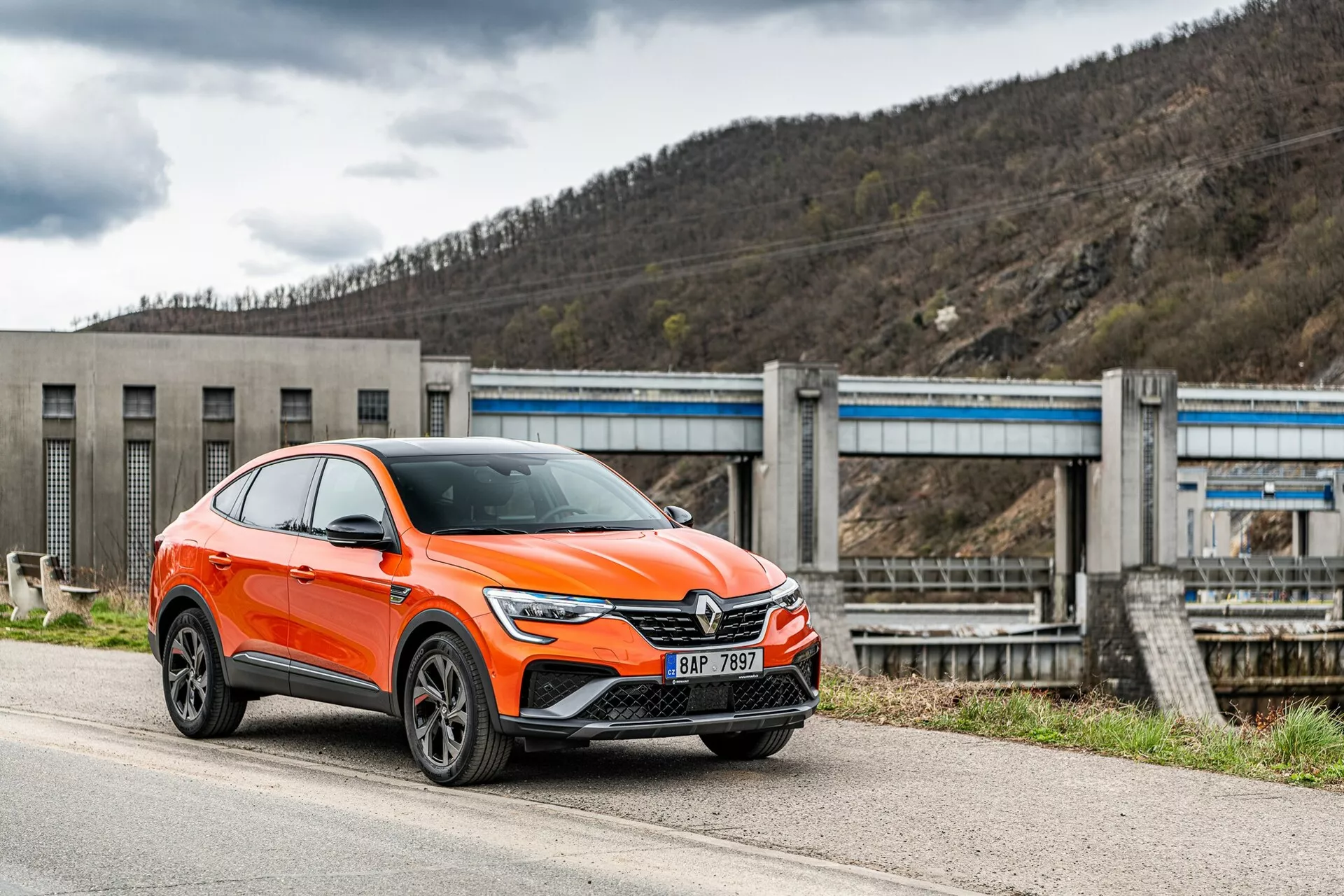En modern, klart orange Renault Arkana 2021 crossover SUV är parkerad vid sidan av en väg, fotograferad från en front-sida vinkel som fokuserar på förarsidan. Bilden visar bilens frontgaller med ett stort emblem, slanka vinklade LED-strålkastare, dagsljusljus, luftintag och eventuellt sensorer för förarassistent på den nedre stötfångaren, uttalade hjulhus med flerspakiga lättmetallfälgar och speglar med integrerade blinkers. Fordonet, klassificerat som en SUV, har en förhöjd hållning med ökad markfrigång och en sluttande taklinje i coupé-stil. I bakgrunden syns en infrastruktur som liknar en damm eller en slussport, vilket placerar bilen i en industriell eller lantlig miljö. Bilden är tagen från en något låg vinkel, vilket betonar bilens höjd och hållning.