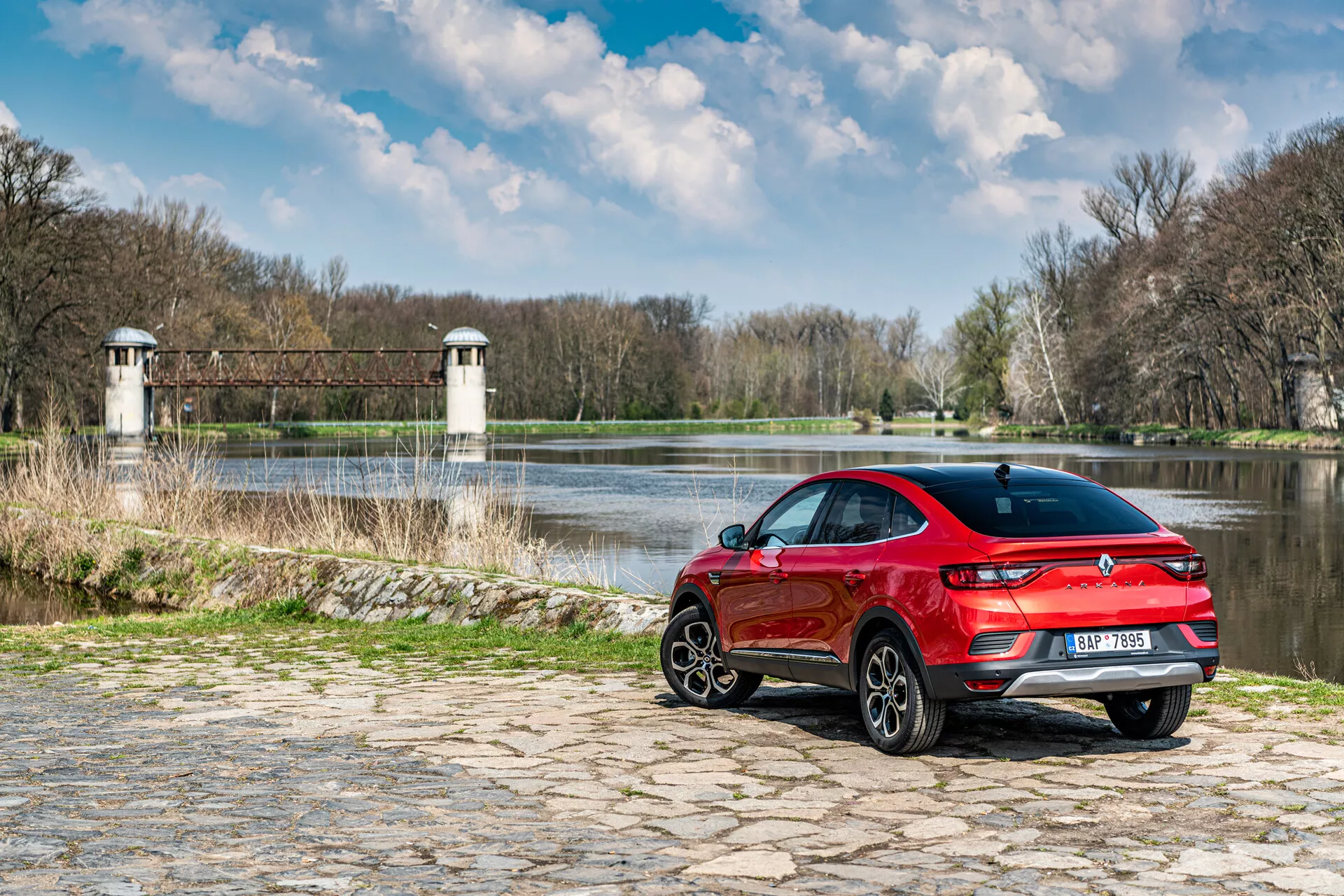 Renault Arkana 2021 parkerad på kullersten nära vatten med naturlig bakgrund, visar bakifrån tre fjärdedels vy, sportig röd design och LED-bakljus
