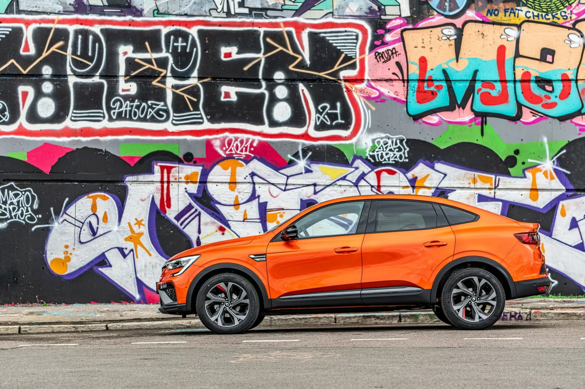 Bilden visar en livfull orange Renault Arkana 2021 crossover SUV parkerad parallellt med en gata med en färgglad graffitivägg i bakgrunden. Fotot fångat i sidovyvinkel framhäver fordonets sportiga och moderna design med synliga förarens sida fram- och bakdörrar, sidorutor, sidospeglar och sluttande taklinje. De stylade lättmetallfälgarna med lågprofildäck antyder fokus på prestanda på vägen. Nyckellösa ingångshandtag på framdörrarna antyder ett nyckellöst inträdes- och startssystem. Bilden är tagen i dagsljus och betonar kontrasten mellan den orange karossen och graffitin och visar bilens välunderhållna, skarpa kaross i en ungdomlig, urban miljö.