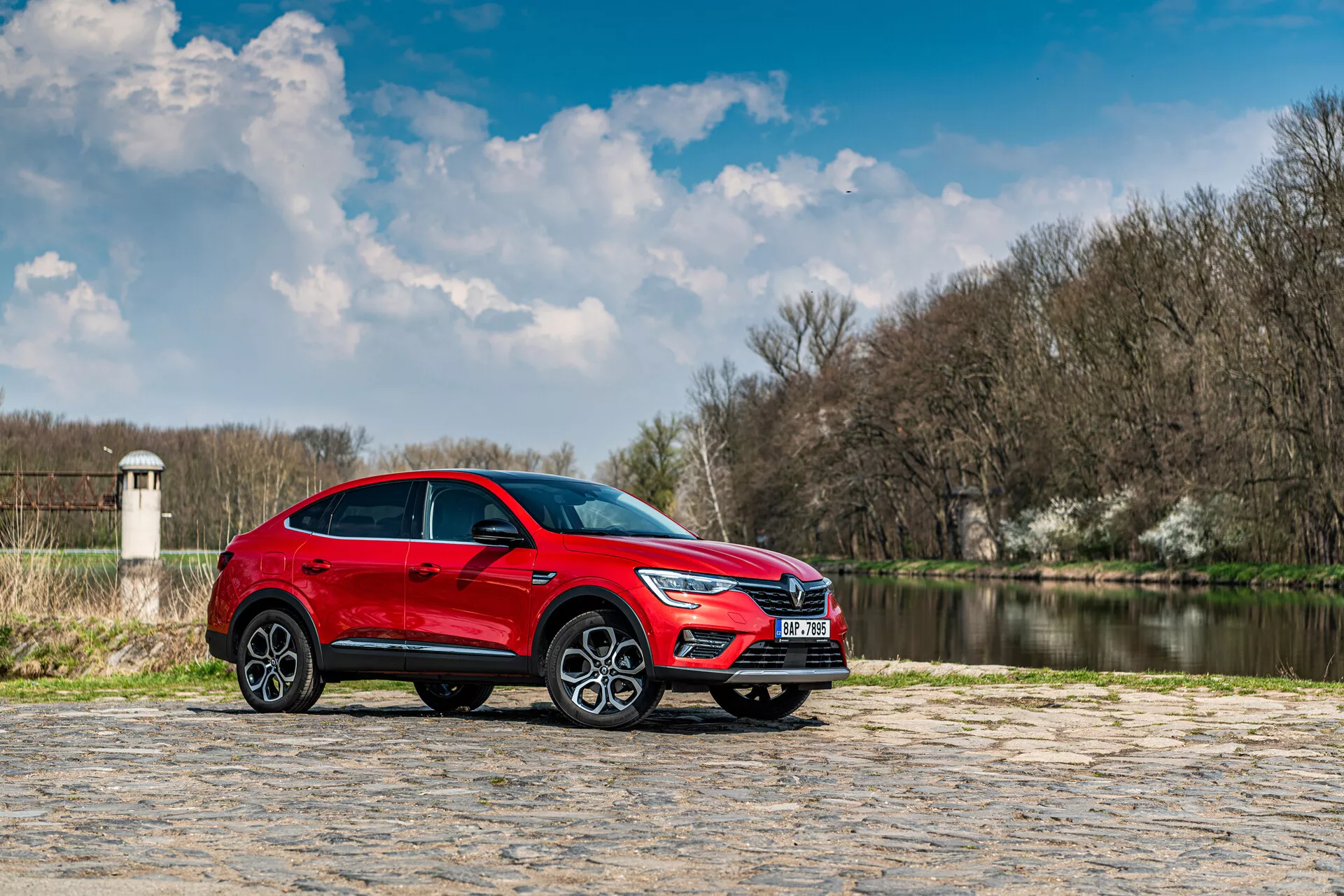 Röd SUV Renault Arkana 2021 parkerad på stenbeläggning med vatten och träd i bakgrunden under delvis molnig himmel, sidoprofil framhäver dess eleganta design och dynamiska funktioner