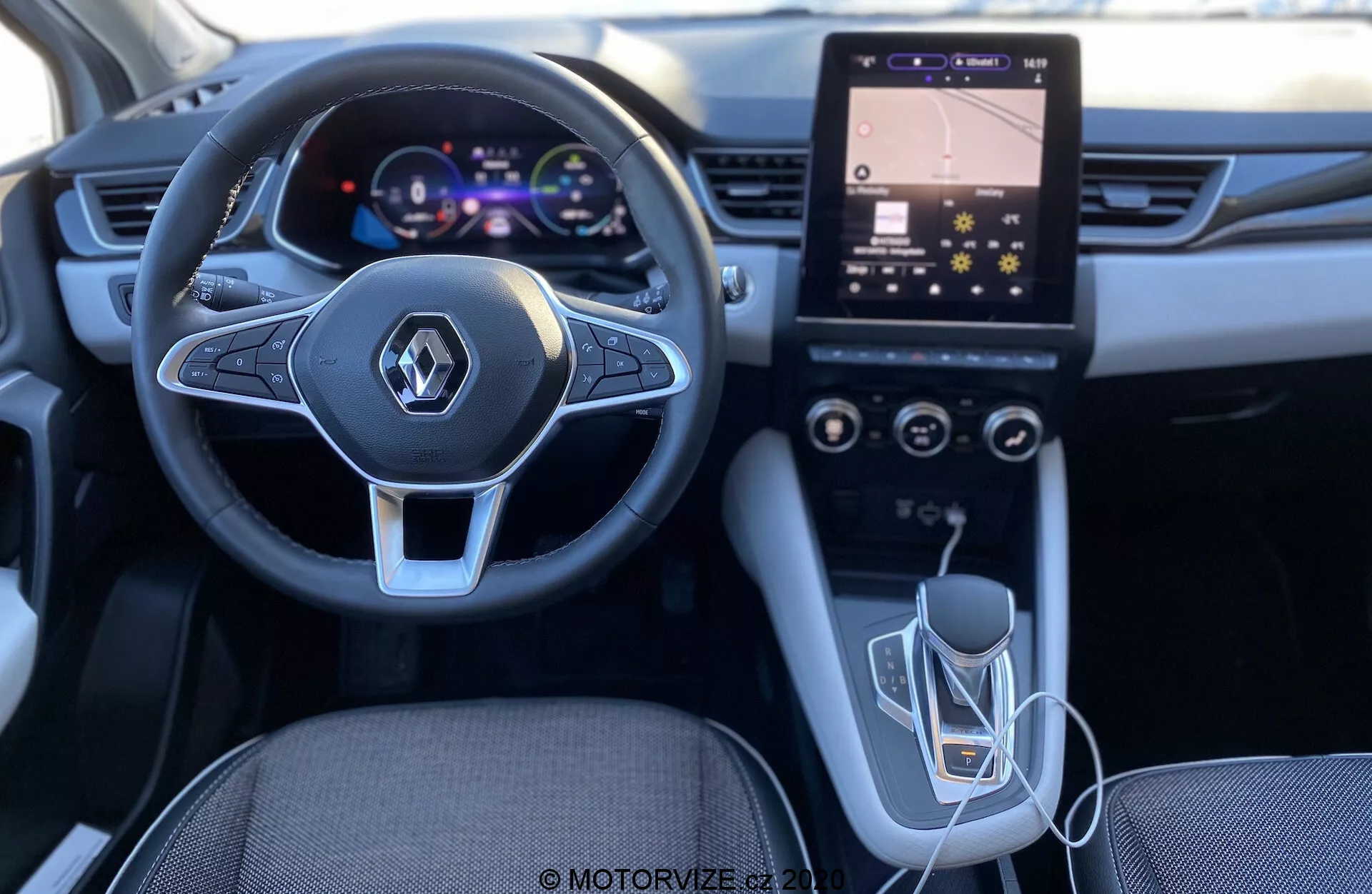 Interiörbild av Renault Captur 2020 från förarens perspektiv som framhäver den moderna designade ratten med integrerade multimedia kontroller, digitalt instrumentkluster, vertikalt orienterad pekskärms infotainmentsystem med HVAC kontroller, automatisk växelspak och USB-anslutning. Belysningen och kvaliteten tyder på en välutformad och tekniskt avancerad interiör.
