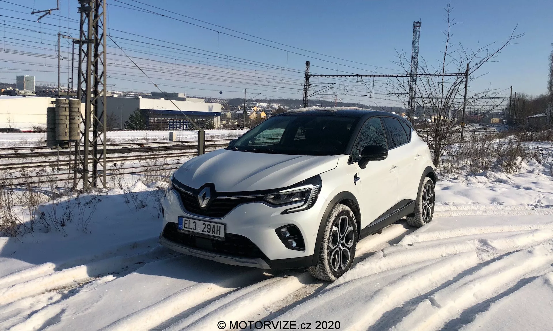 Bilden visar en vit Renault Captur 2020, en kompakt SUV parkerad vid en snötäckt vägkant, fotograferad från en trekvarts framvinkel. Fordonet har distinkta C-formade LED-dagljus, projektorstrålkastare, en svart accenterad framgrill med ett framträdande Renault-emblem och flerspokes legeringshjul. Anmärkningsvärda designelement inkluderar plastbeklädnader längs de nedre kanterna och hjulhusen. Miljön antyder en kall vinterdag med en industri- eller halvurban bakgrund.