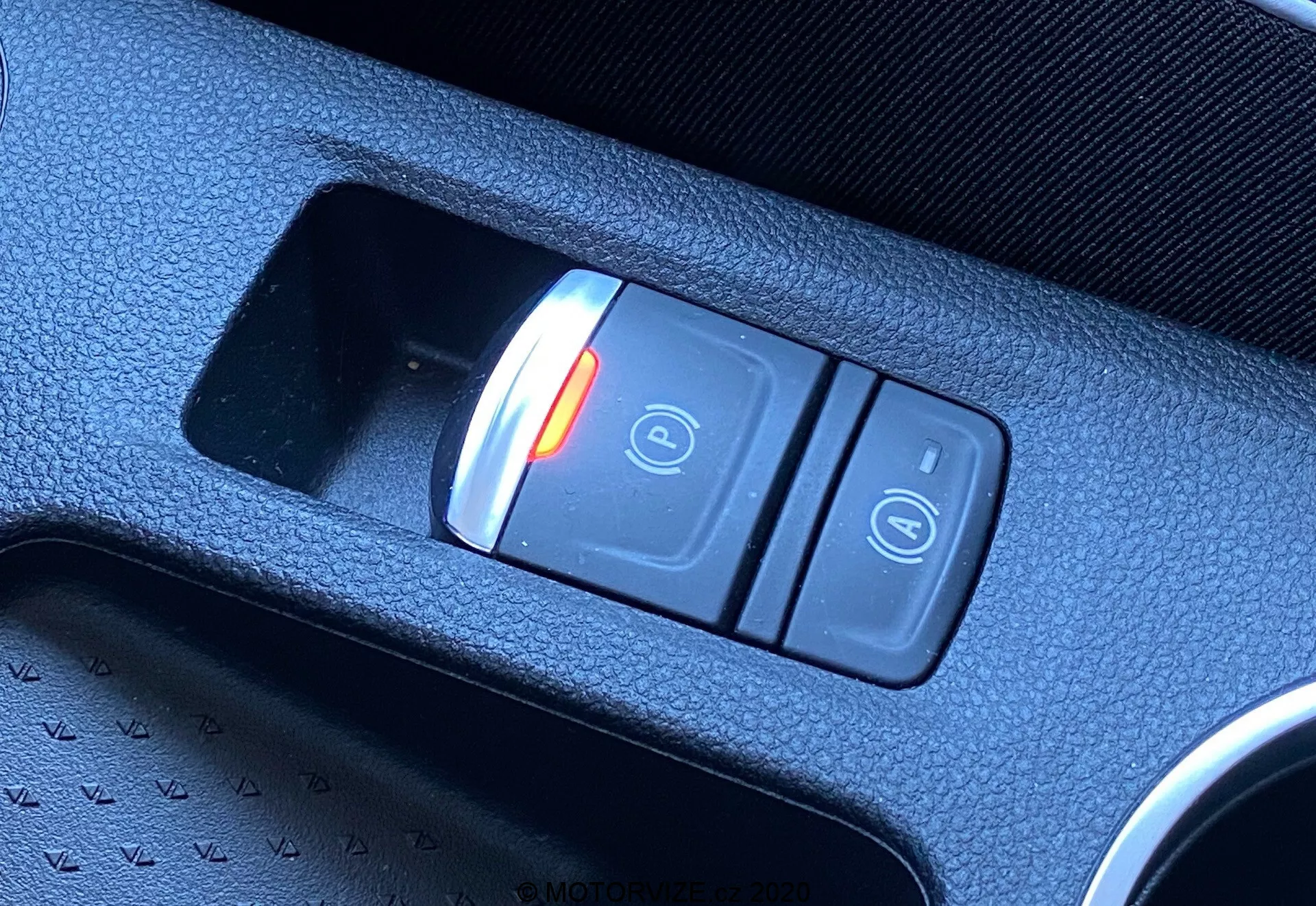 Närbild i diagonal vinkel av den elektroniska parkeringsbromsen i Renault Captur 2020:s interiör. Omkopplaren, placerad i den centrala konsolen, har två knappar: en röd upplyst knapp med en stängd hänglås och 'P'-symbol som indikerar aktiverat läge, och en annan knapp med symbolen för ett öppet hänglås. Omkopplaren är placerad i en strukturerad svart plastkonsol, som framhäver modern fordons teknik.