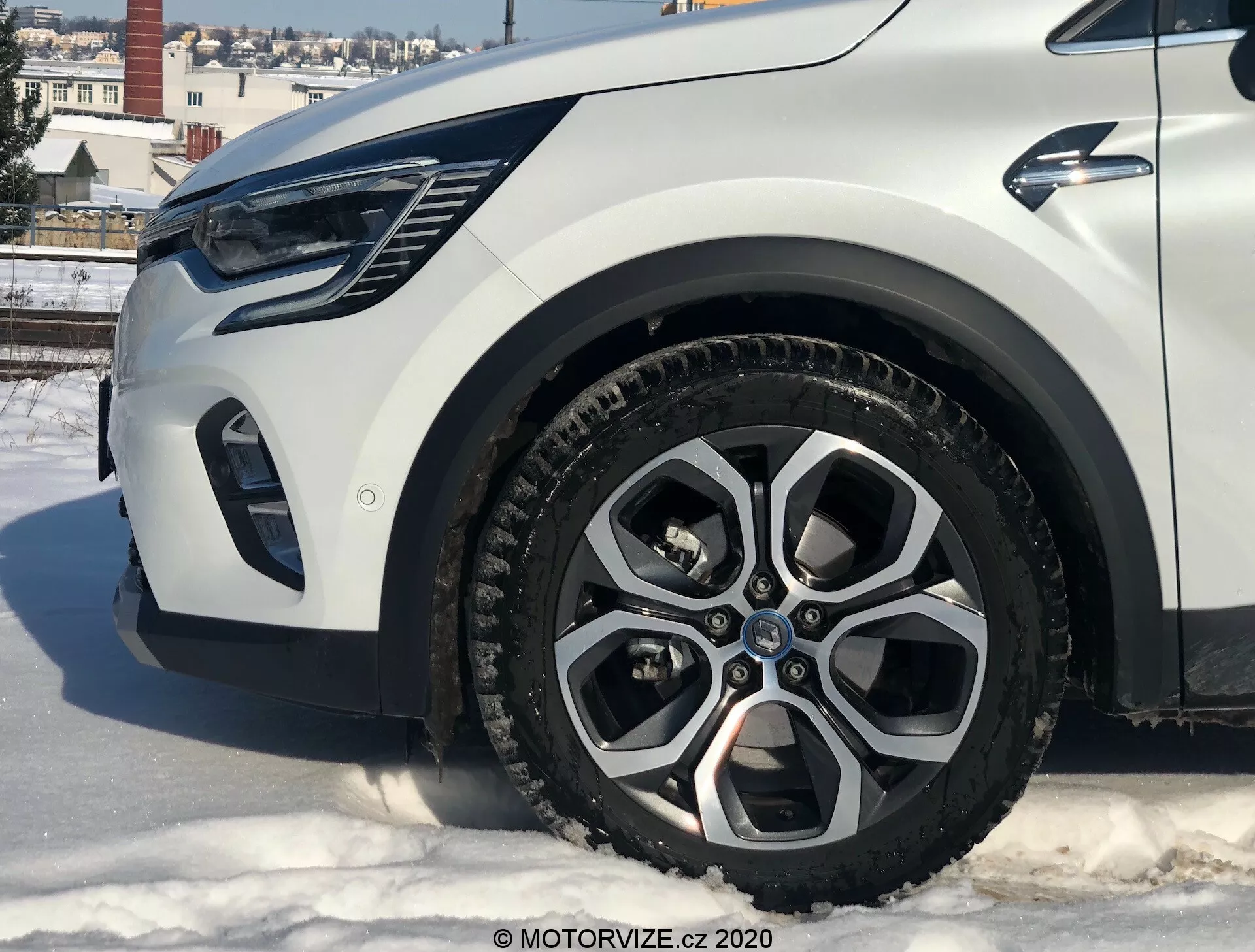 Närbild av en Renault Captur 2020 från låg vinkel som visar vänstra framdelen, med fokus på lättmetallfälgen, skärmbreddaren, stötfångaren och strålkastarnas område. Bilen står på en snötäckt yta i en urban miljö med byggnader och klar himmel.