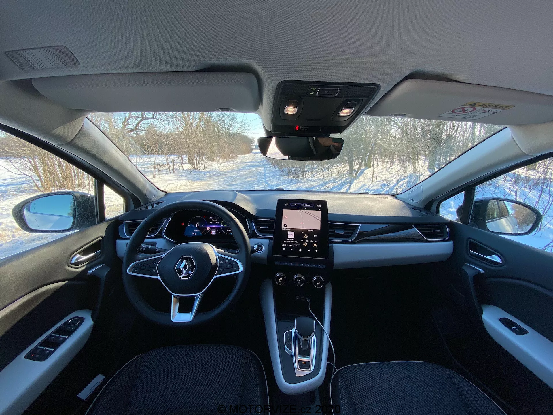 Interiörbild av 2020 Renault Captur från baksätena som visar framkabinen med en treekrad ratt med integrerade knappar, digital instrumentpanel, vertikalt orienterat pekskärmsinfotainmentsystem, modern mittkonsol med minimala fysiska kontroller, elegant växelväljare, ventilationsöppningar för HVAC, premium material inklusive läder och metalliska accenter, backspegel möjligen med sensorer för ADAS och synlig A-stolpe med fönsterkontroller.