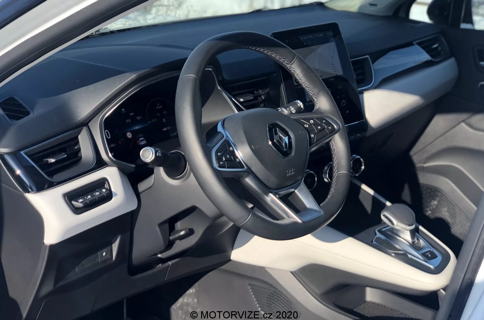 Interiörvy av 2020 Renault Captur från förarens perspektiv, med fokus på ratten med Renault-logotypen, monterade kontroller, digitala instrumentklustret bakom och ett centralplacerat pekskärms-infotainmentsystem. Bilden framhäver en blandning av syntetiska läder- och plastmaterial med metalliska accenter, automatisk växellåda, klimatanläggning och justeringar av fönster och speglar.