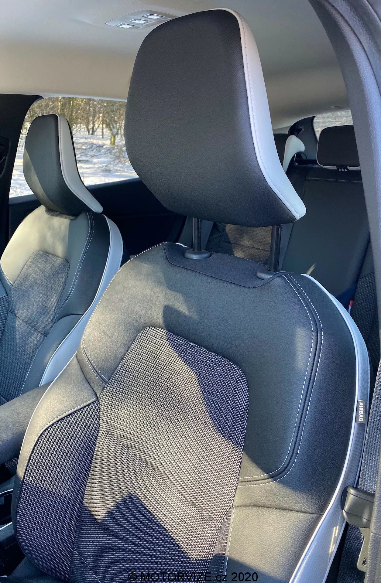 Interiören i en Renault Captur 2020, som visar passagerar- och förarsätet framifrån en bakre, något off-center vinkel. Den övre delen av sätena inkluderar integrerade nackstöd och synliga ramar för säkerhetsbälten. Sätena har en modern tvåtonig design i mörkgrå och ljusgrå färg, med syntetläderkanter och detaljerade sömmar som framhäver hantverk och stil.