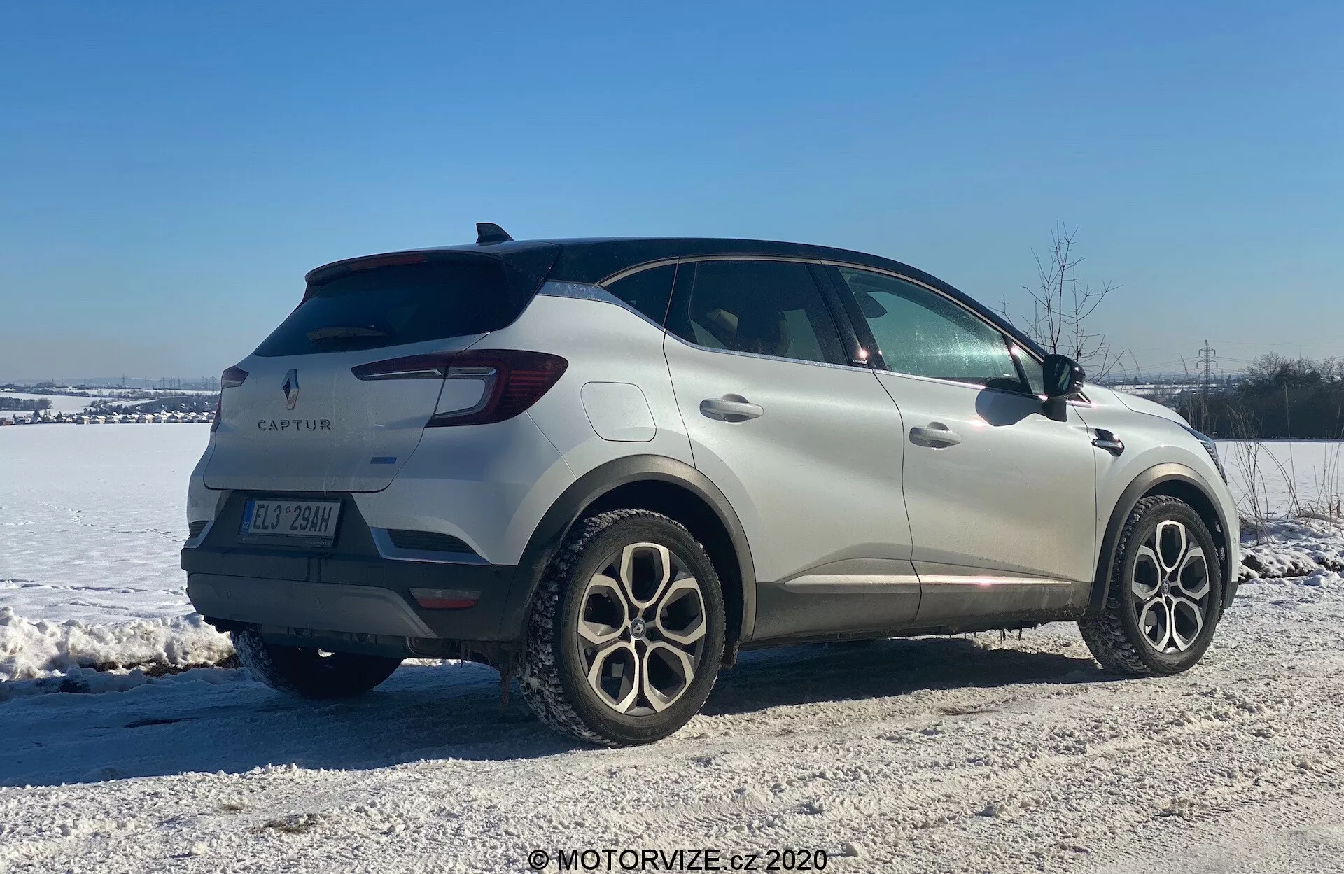 Bild av Renault Captur 2020, en kompakt SUV, i en vintermiljö. Visad från trekvarts bakre vy, bilen har en elegant design med mjuka linjer och LED-bakljus. Tillverkarens emblem och modellnamnet är synliga tillsammans med flerspakiga lättmetallfälgar, bakre parkeringssensorer och sidospeglar med inbyggda blinkers. Parkerad på en snötäckt grusig yta, antyder bilden skicklighet i olika terränger och vinterförhållanden.