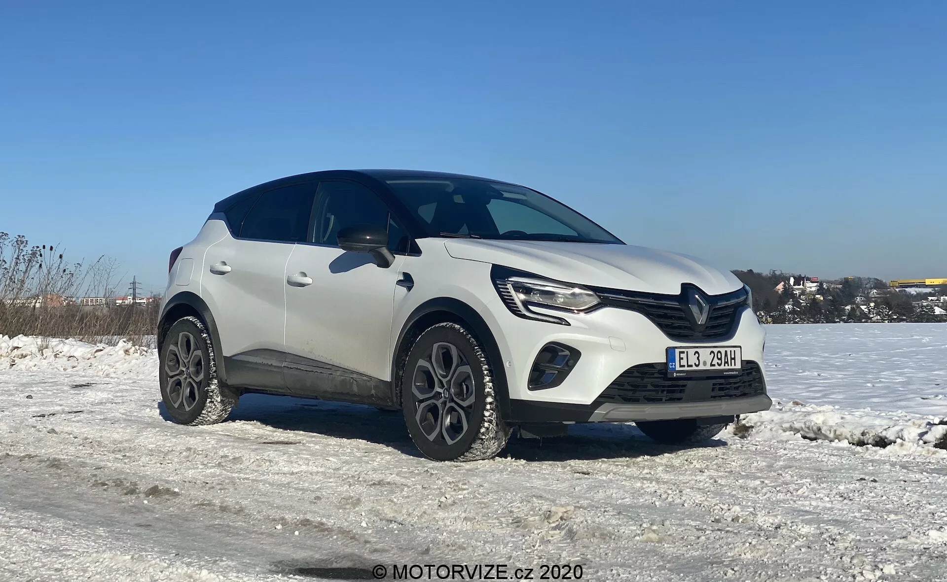 Trekvartsvy av en vit Renault Captur 2020 kompakt crossover SUV i en vintermiljö, med snötäckt mark och en klarblå himmel. Bilden lyfter fram fordonets vänstra framsida och en del av dess sidoprofil, tagen från en något lägre vinkel som framhäver hjulen och den nedre halvan. Bilens moderna design visar karakteristiska linjer på motorhuven, eleganta LED-strålkastare med dagsljus och en framträdande främre grill. Unika legeringshjul och plastklädsel runt hjulbågarna ger ett robust utseende. Skuggorna under bilen tyder på att fotot togs mitt på dagen eller tidig eftermiddag i naturligt dagsljus.