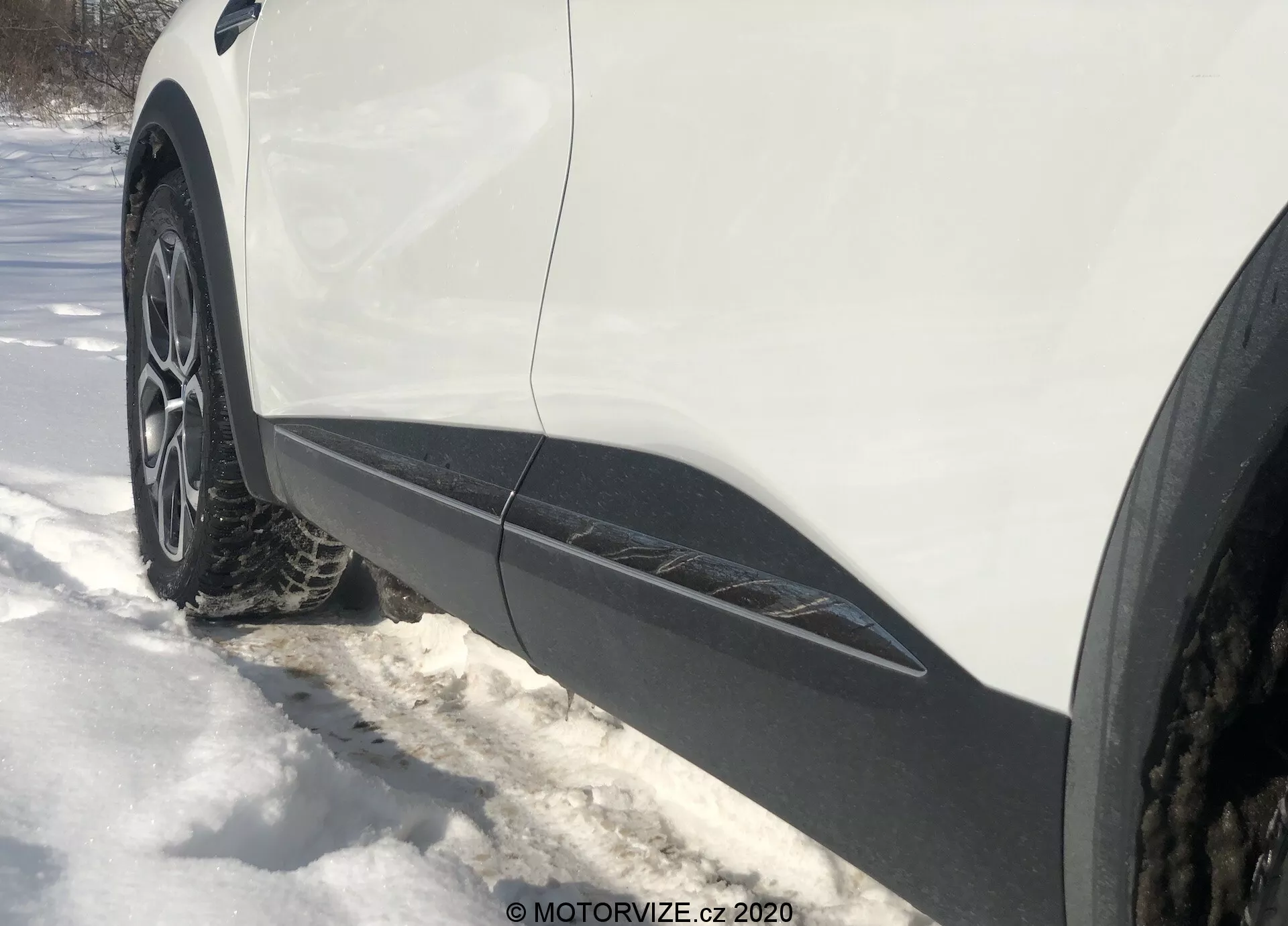 Närbild, sned sidovy från marknivå av vänster framhjul och skärm på en Renault Captur 2020. Den flerekrade legeringsfälgen med vinter- eller året runt-däck omges av snö. Den vita karossen med svart list längs underkanten indikerar en crossover eller SUV. Belysningen antyder en solig dag med synliga skuggor.