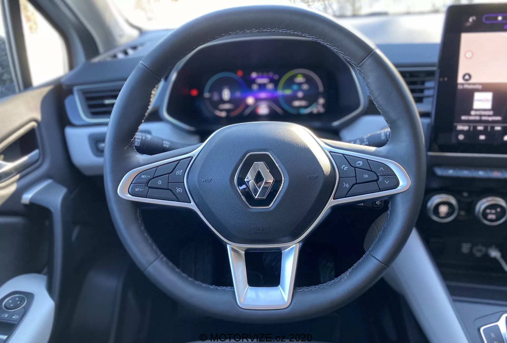 Närbild i första person från förarsätet i en Renault Captur 2020, fokuserad på den treekrade multifunktionsratten med läderklädsel och silverdetaljer samt Renault-logotyp, tillsammans med ett färgglatt digitalt instrumentkluster som visar hastighetsmätare och fordonsdetaljer, med en delvis synlig infotainmentskärm i bakgrunden.