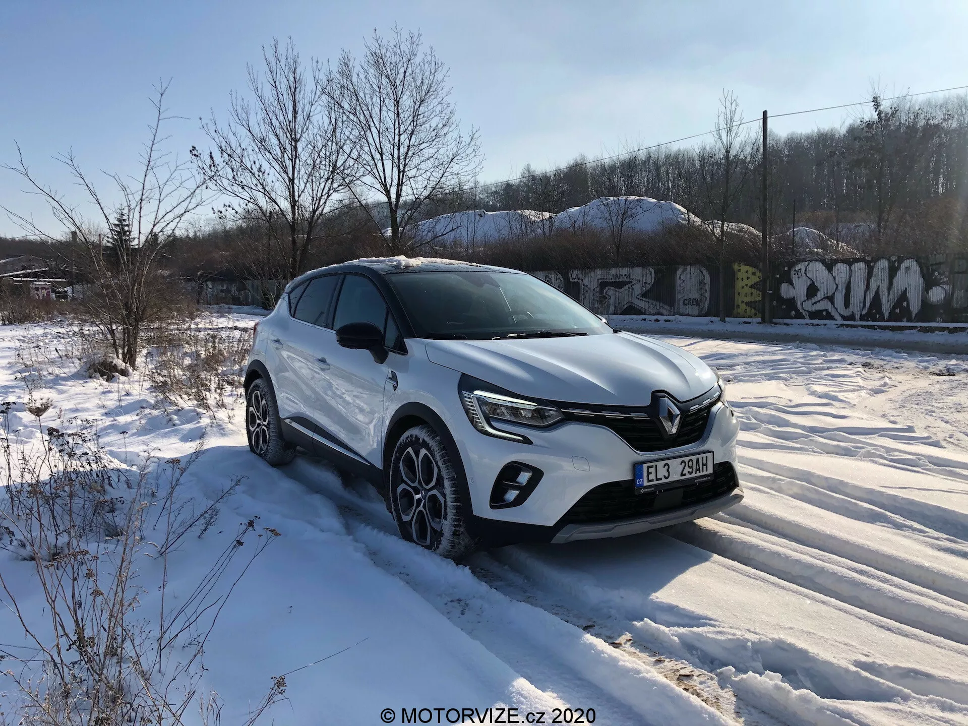 En vit Renault Captur 2020 crossover SUV är parkerad på en snötäckt mark med växtlighet och graffiti-täckta strukturer i bakgrunden, under en mestadels klar himmel med några moln. Bilden är tagen från en trekvart framvinkel fokuserad på förarsidan, som visar frontgrillen med ett kromemblem och moderna LED-strålkastare. SUV:en har främre LED-dagljus, dimljusfångare, karaktärslinjer på motorhuven, plastbeklädnad runt hjulbågarna, integrerade blinkers i sidospeglarna, mångekeleggsfälgar med vinterdäck och en stark axellinje. Den förhöjda ställningen antyder högre markfrigång, lämplig för olika terränger.