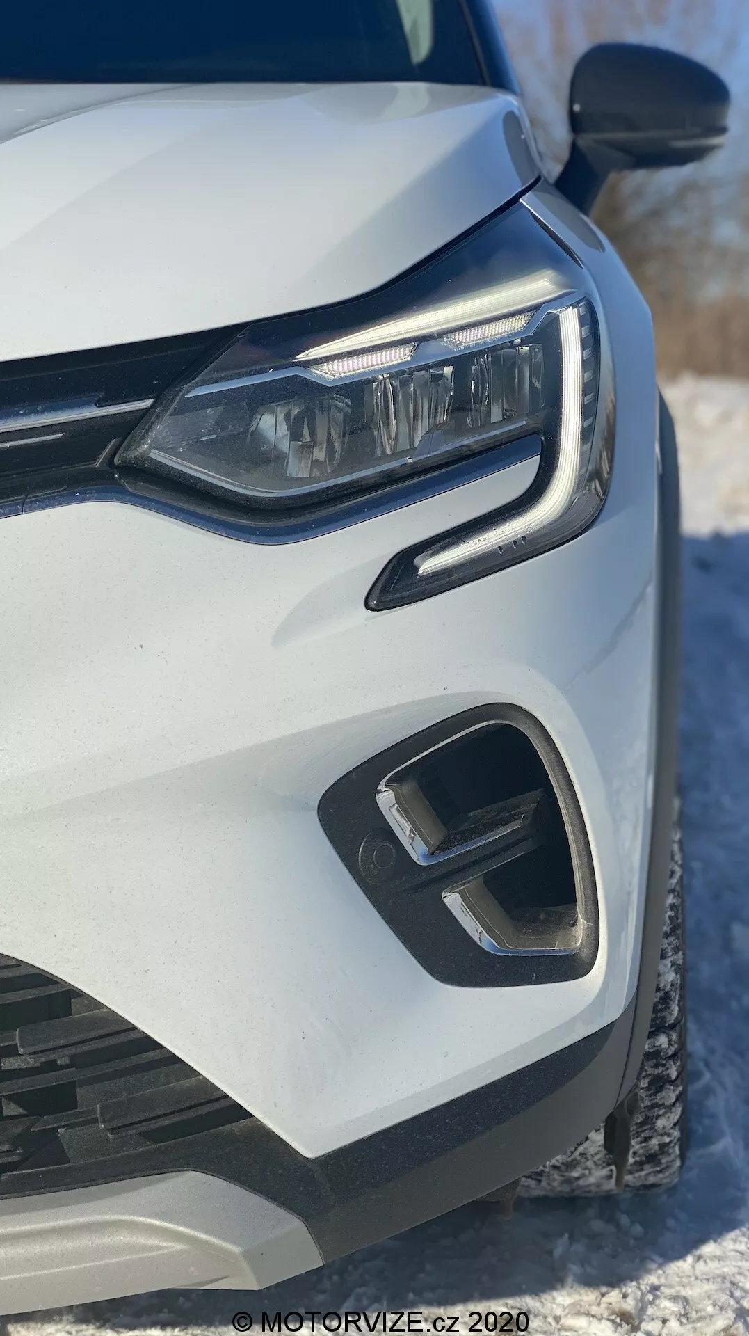 Närbild på vänster framdel av en vit Renault Captur 2020 med fokus på moderna LED-strålkastare, geometriska främre stötfångaren och grilldelar, tagen från en låg framvinkel med snö i bakgrunden