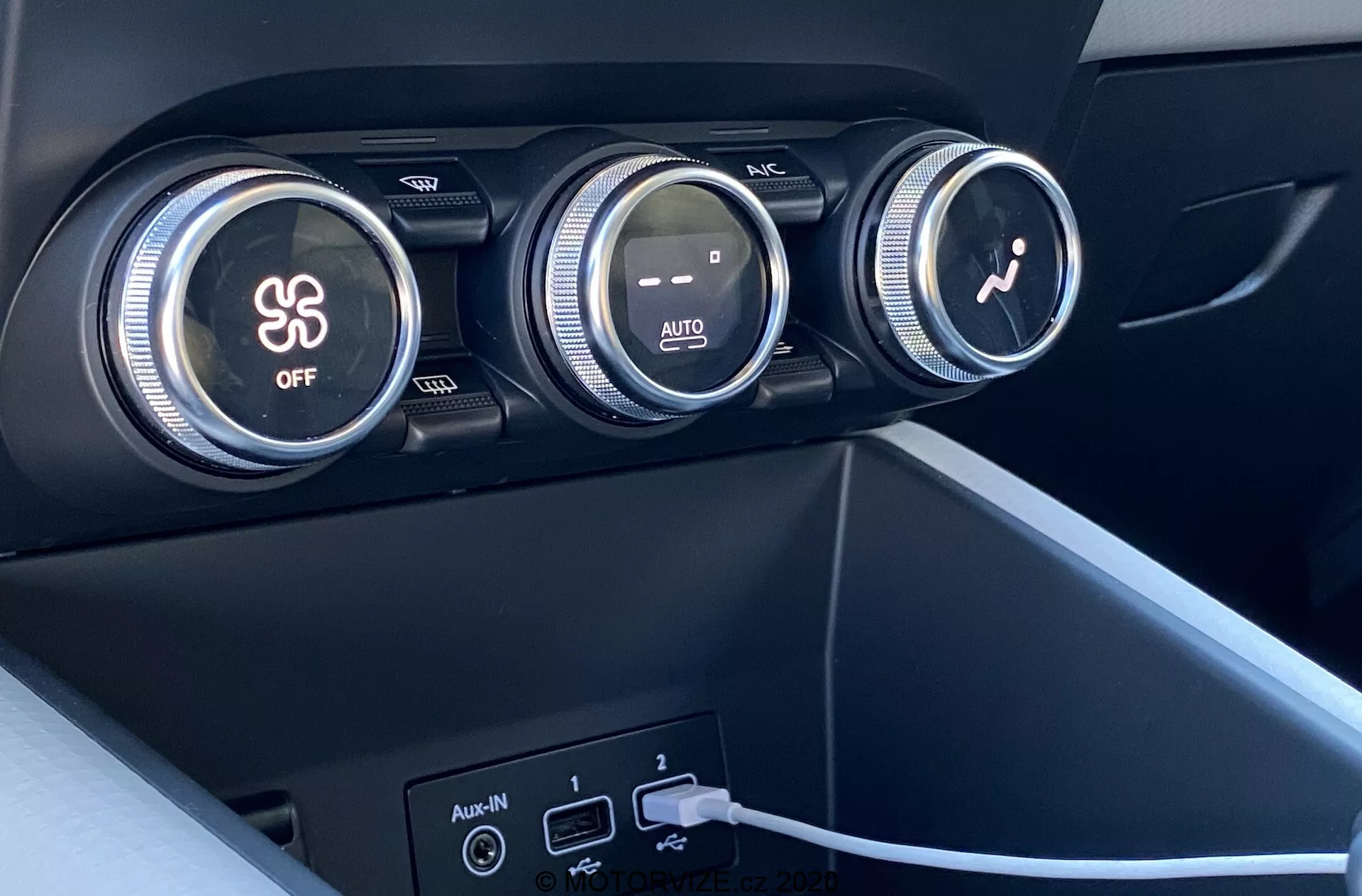 Närbild av interiören i Renault Captur 2020 med fokus på klimatanläggningen. Bilden visar tre stora reglage på instrumentbrädan. Det första reglaget styr luftkonditioneringen, markerat med en snöflingesymbol och en 'OFF'-knapp. Det mellersta reglaget reglerar temperaturen med röda och blå färggraderingar och 'AUTO'-indikering. Det tredje reglaget med en fläktikon justerar fläkthastigheten. Under reglagen finns två USB-portar och en AUX-ingång, med en kabel inkopplad i en av USB-portarna. Materialen är mörk plast eller syntet med matt finish och en modern minimalistisk design.