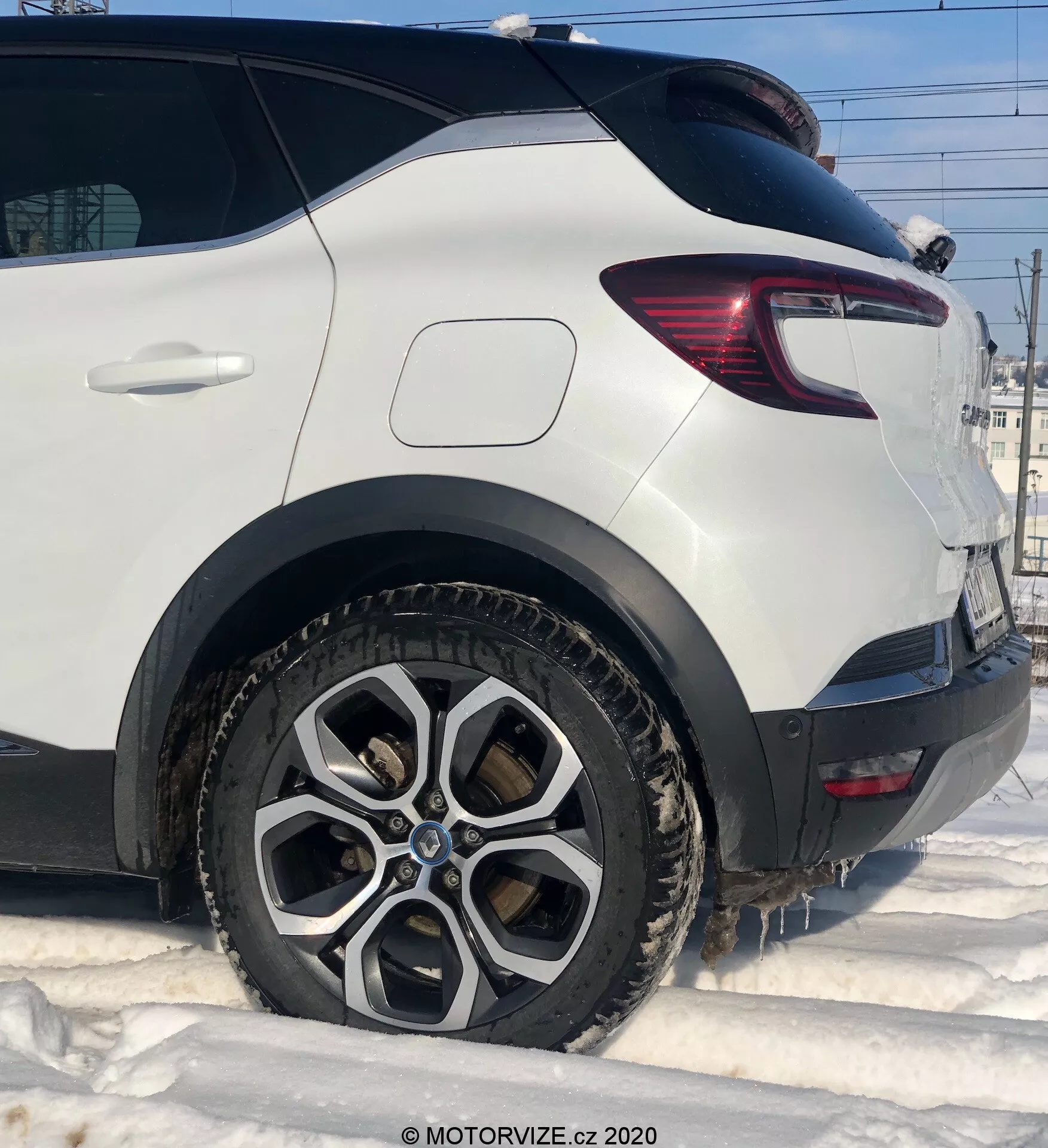 Detaljerad närbild på en vit Renault Captur 2020 som visar den bakre passagerarsidan från låg vinkel. Detaljer inkluderar det bakre hjulet med en flerekrad legeringsdesign, smutsigt av lera och snö, som antyder vinterförhållanden. Bilden fångar den rena bakre skärmen ovanför hjulbågen, den moderna baklyktan med röda och klara LED-sektioner och robust plastbeklädnad bakom hjulet. Det sluttande bakfönstret bidrar till fordonets aerodynamiska form.