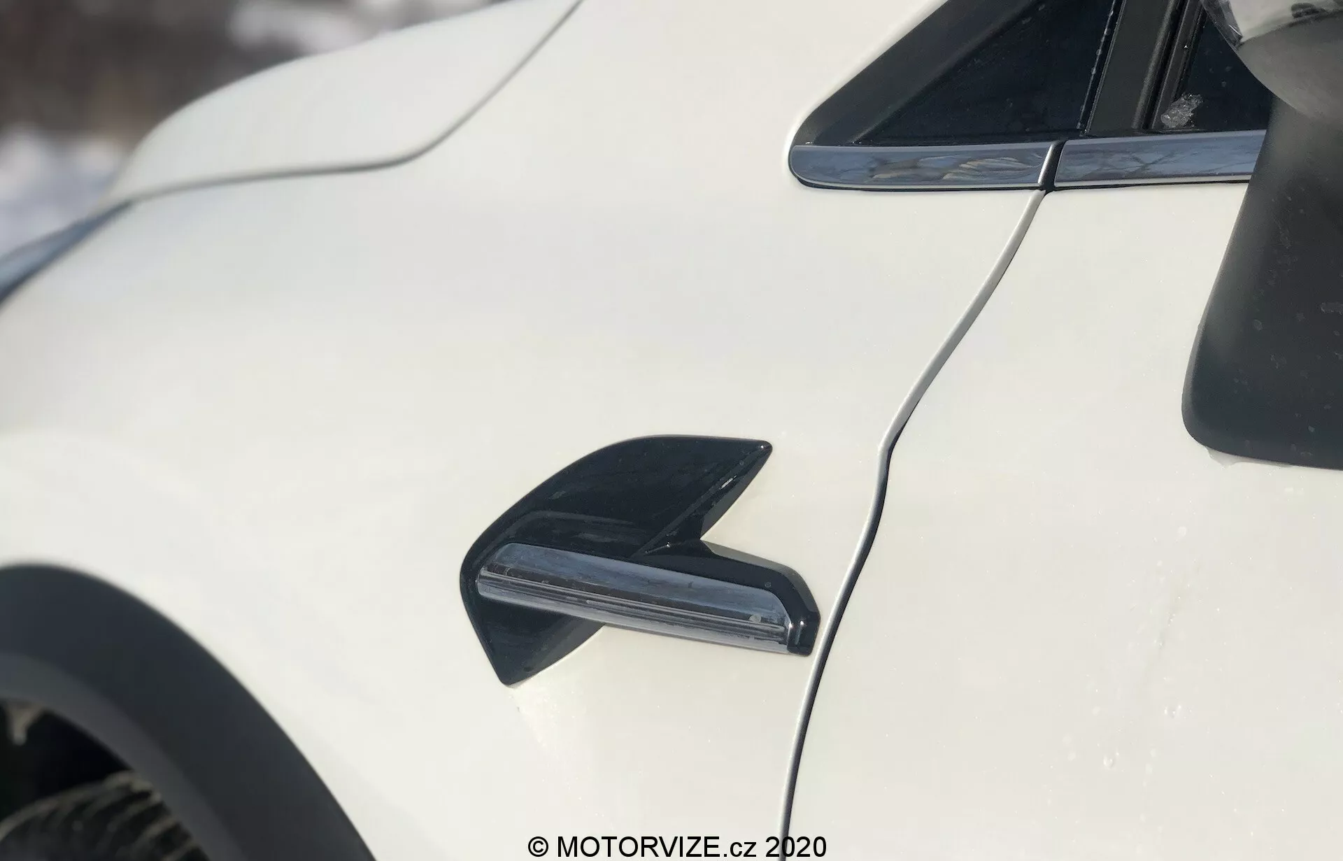 Närbild av utsidan av Renault Captur 2020, med fokus på sidan av det vita fordonet. Bilden visar en svart, strömlinjeformad hajfensantenn i övre vänstra hörnet, utformad för radiomottagning och aerodynamik. Centrerad under antennen framhävs en svart luftventil eller luftintag, troligen för bromskylning eller förbättrad luftcirkulation. Bilens aerodynamiska taklinje böjer sig bakåt, med en glansig vit lack som reflekterar ljuset. Bakgrunden är något suddig, vilket indikerar en snöig utomhusmiljö.