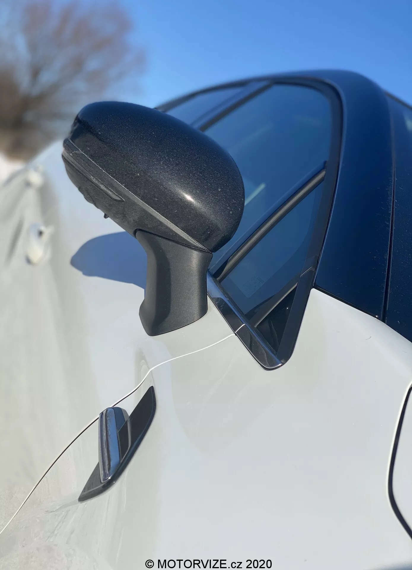 Närbild av Renault Captur 2020:s sidospegelmontering framifrån och bakåt, med fokus på det svarta texturerade plastfäste med integrerad blinkers och som reflekterar den blå himlen. Den eleganta mörkfärgade karosspanelen nedanför och den möjliga döda vinkelspegeln indikerar en modern design och avancerade funktioner.