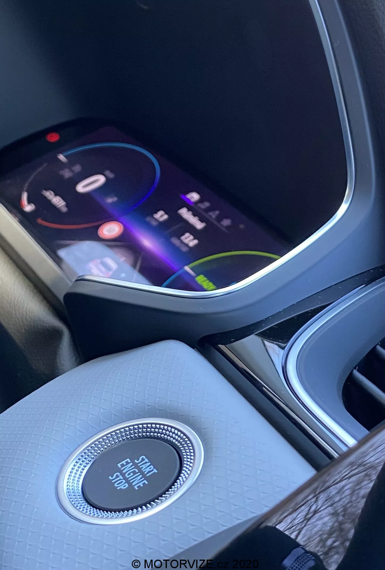 Interiör av en Renault Captur 2020 med fokus på mittkonsolen och instrumentbrädan ur ett framsätesperspektiv. Den livliga infotainmentskärmen visar ett färgglatt gränssnitt som antyder avancerade multimediefunktioner, inklusive navigation, medieuppspelning och anslutningsalternativ. Under skärmen finns en rund kontrollknapp med kromfinish märkt 'Engine Start Stop' som framhäver nyckelfritt startsystem. Mittkonsolen har en högkvalitativ texturerad finish som förbättrar interiörens atmosfär. Till vänster syns delvis en mugghållare eller förvaringsfack.