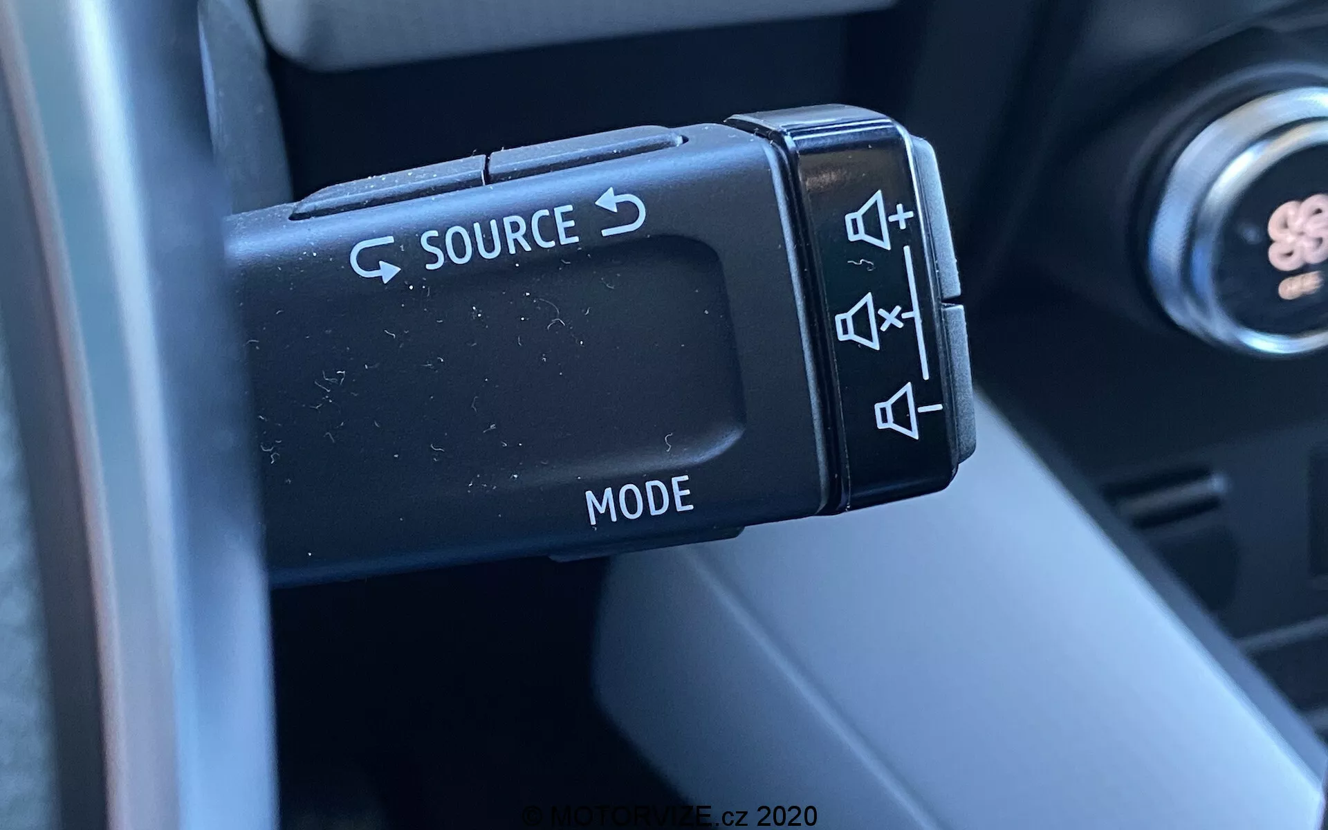 Närbild av de rattmonterade ljudkontrollerna i en Renault Captur 2020, med fokus på vänster kontrollspak med knappar för att byta källa (SOURCE), justera volymen, hoppa över spår och välja läge (MODE). Fotografiet fångar detaljerna på de matta plasthandtagen och deras ergonomiska design, med en del av rattstången och bilens interiör i suddig bakgrund, vilket framhäver funktionalitet och bekvämlighet för föraren.