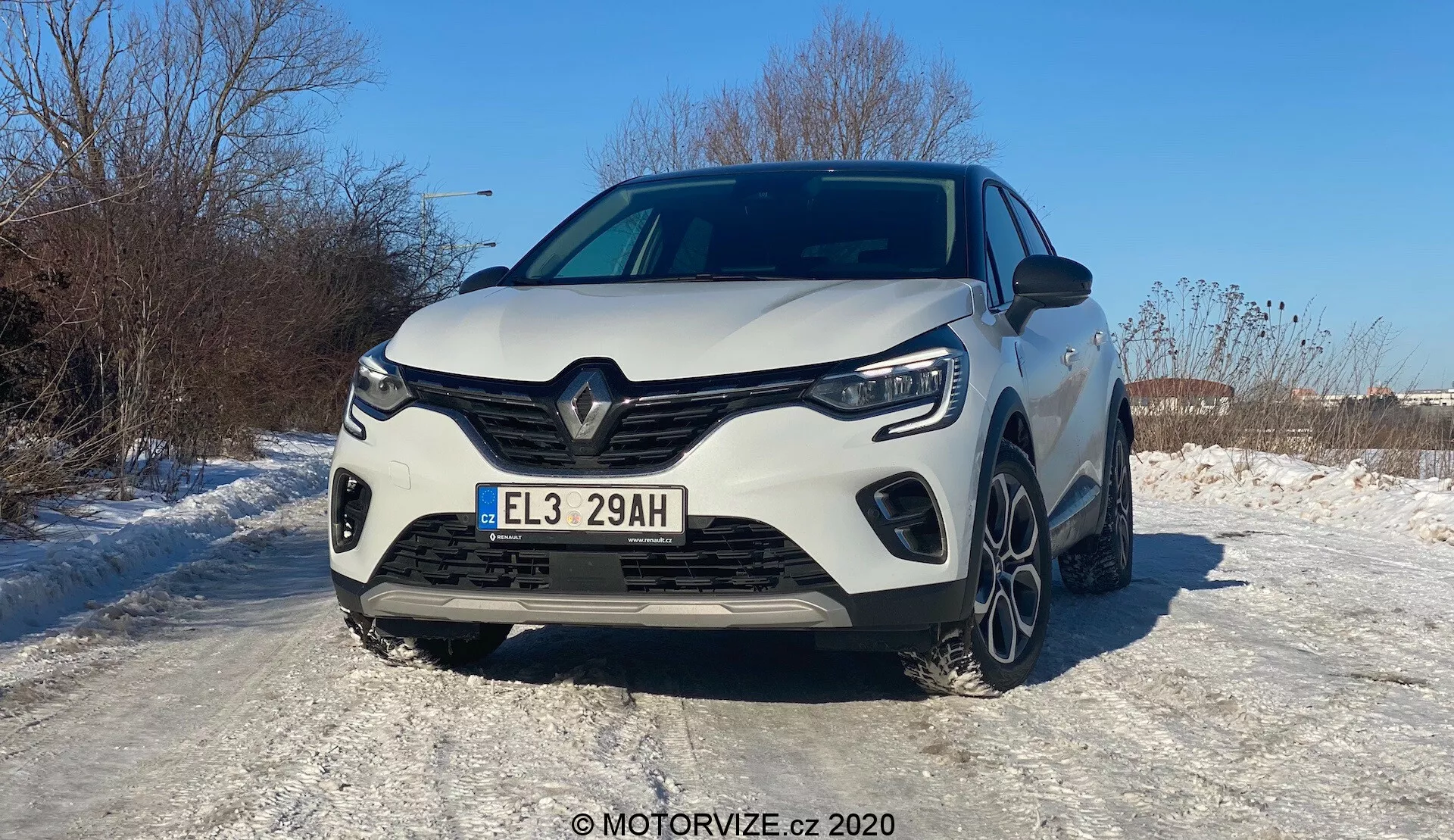 Frontal vy av en vit 2020 Renault Captur, fotograferad i en vinkel som något favoriserar den vänstra sidan. Den moderna bilen visar en utmärkande Renault-grilldesign med en stor central logotyp, skarpa linjer och skulpterade konturer. Hela frontgrillen, LED-strålkastarna och de vinklade dagsljusen syns tydligt. Den sportiga stötfångaren har framstående sidoluftintag och en centrala sektion som troligen är för kylning. Motorhuven har upphöjda linjer för ett muskulöst utseende. En del av framhjulen och multi-ekrade lättmetallfälgar är synliga på den snötäckta ytan, vilket indikerar vinterförhållanden. Den europeiska registreringsskylten och den glänsande lacken som reflekterar solljus tyder på en solig, klar dag. Avancerade förarassistanssystem, LED-belysning och hybrid/elektrisk drivlina kan antas från 'E' på skylten.