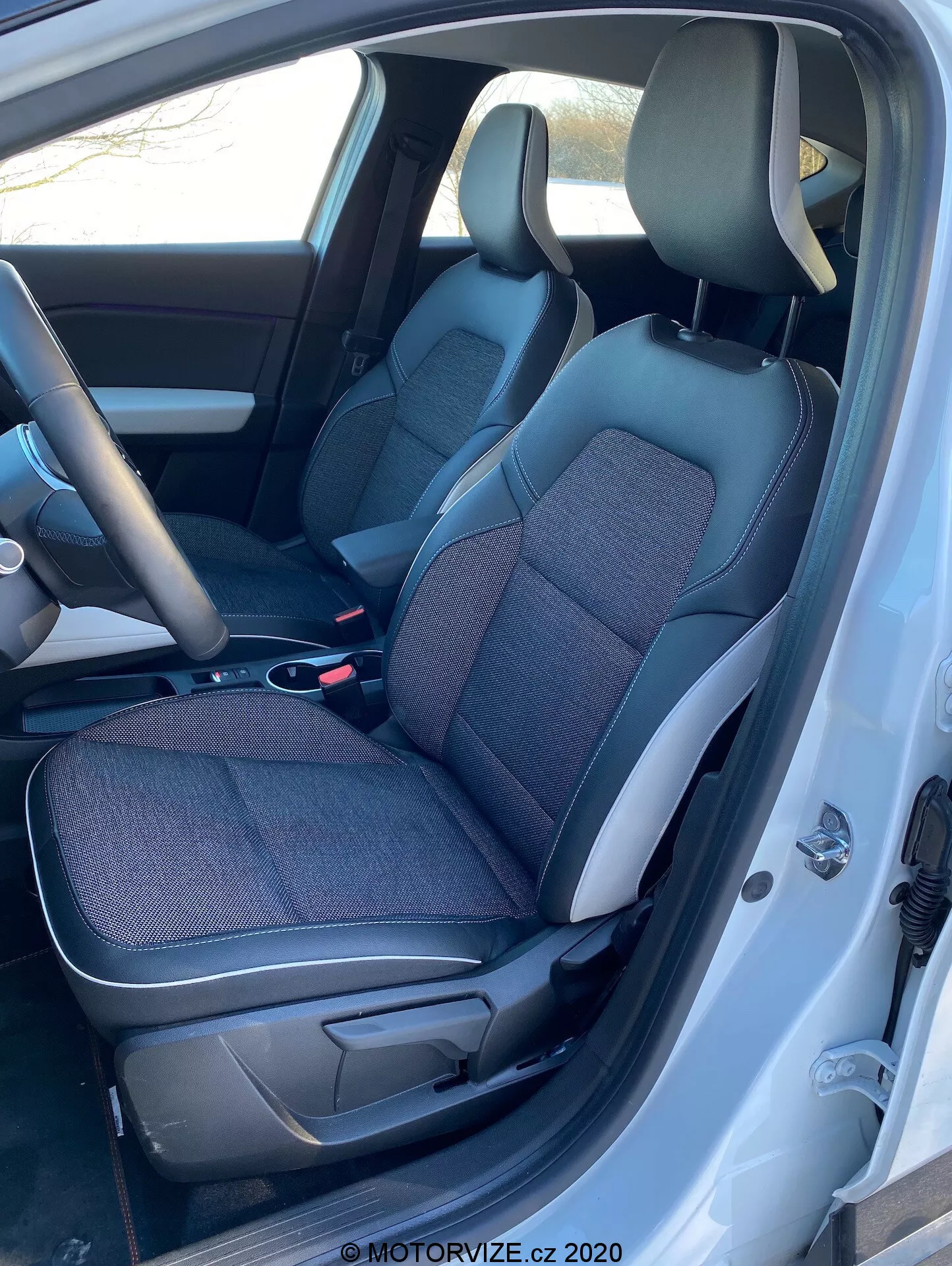 Interiörsbild av Renault Captur 2020 från vänster sida med förardörren öppen, visar svarta och grå tyg- och lädersäten, mittkonsol, ratt och en del av instrumentbrädan.