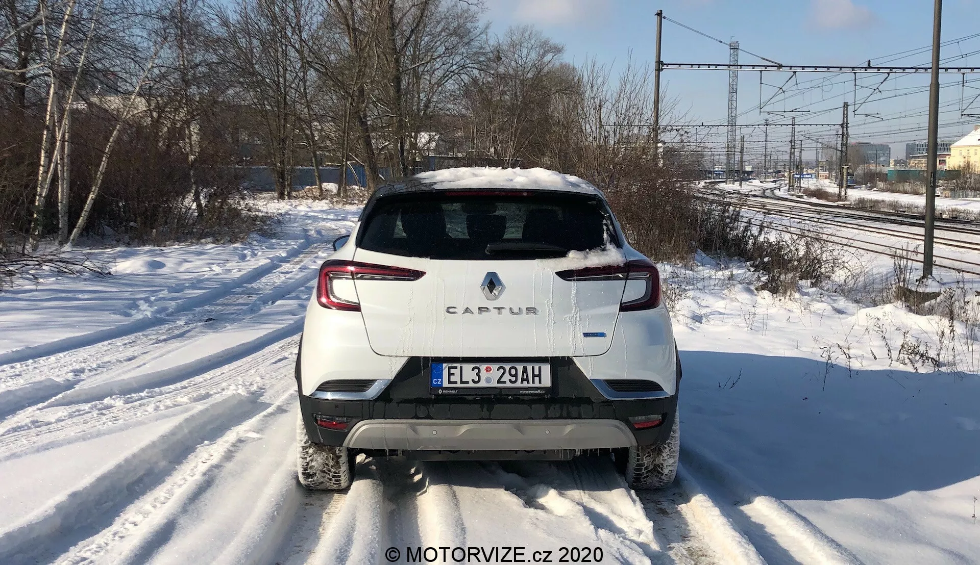 Bakåtvänd bild av en Renault Captur 2020 på en snötäckt väg. Bilden lyfter fram den distinkta bakre designen, inklusive det tvåfärgade färgschemat med en vit karosseri och kontrasterande tak, modern bakre LED-belysning, europeisk stilregistreringsskylt, bakrutetorkare och ett enda avgasrör på höger sida. Fotot fångar kraftledningar och trädlinjer i bakgrunden under dagsljus, vilket antyder en förorts- eller halvlandsbygdsmiljö.