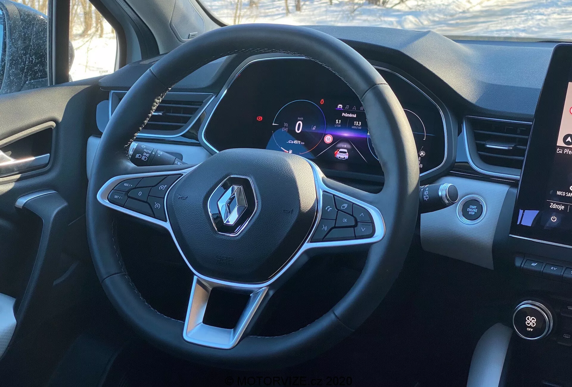 Interiörvy av 2020 Renault Captur från förarens perspektiv, med fokus på den sportiga, plattbottnade multifunktionsratten med Renault-logotypen och kontrollknappar för ljud- och farthållningsfunktioner. Den digitala instrumentpanelen visar en stillastående hastighet på 0 km/h bredvid varvräknaren, vilket indikerar modern teknik och anpassningsbar förarinformation. Till höger syns en del av den vertikala pekskärms-infotainmentsystemet som visar temperaturreglage och medieinformation, i dagsljusförhållanden som ger en realistisk förarvy.